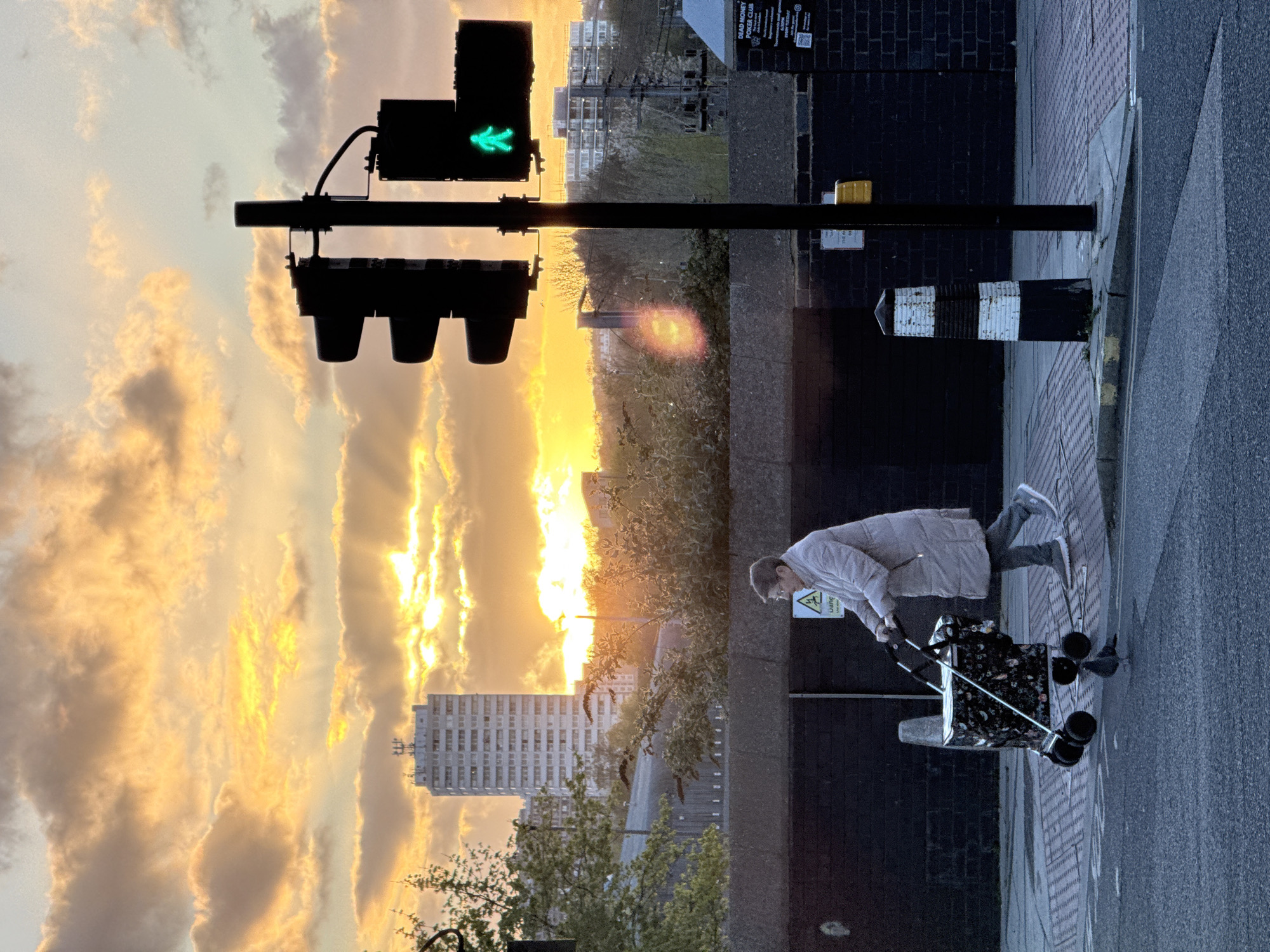A pedestrian walks along a London sidewalk at sunset. The traffic light is green. A tall building can be seen in the background. The pedestrian is pulling a shopping cart.