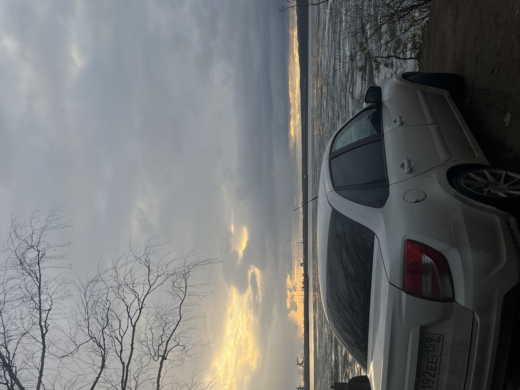 A white sedan is parked on a dirt bank next to a large body of water, viewed from a slightly elevated angle. The image is originally oriented vertically but depicts a horizontal landscape. The car, a four-door sedan, is predominantly white and appears somewhat dirty, particularly on its rear bumper and wheel arch, suggesting travel through muddy conditions. The car's rear left side is prominent, with a visible taillight and a Russian license plate that reads "M 192EE 159 RUS".
The body of water, likely a river or lake, is partially covered with broken ice or slush, indicating cold weather conditions. Along the distant horizon, a striking band of golden light from the sun illuminates the clouds and reflects on the water's surface, suggesting either sunrise or sunset. Above this glowing horizon, the sky is largely overcast with dense, dark clouds, though patches of lighter sky are visible.
To the left of the frame, several bare, deciduous trees with intricate, leafless branches stand against the cloudy sky. Their skeletal appearance further emphasizes the cold, possibly late autumn or early spring season. The overall scene conveys a sense of serene solitude and the stark beauty of a natural landscape at dawn or dusk in a cold climate, typical of locations like Chaikovsky, Russia. No people are visible in the image.