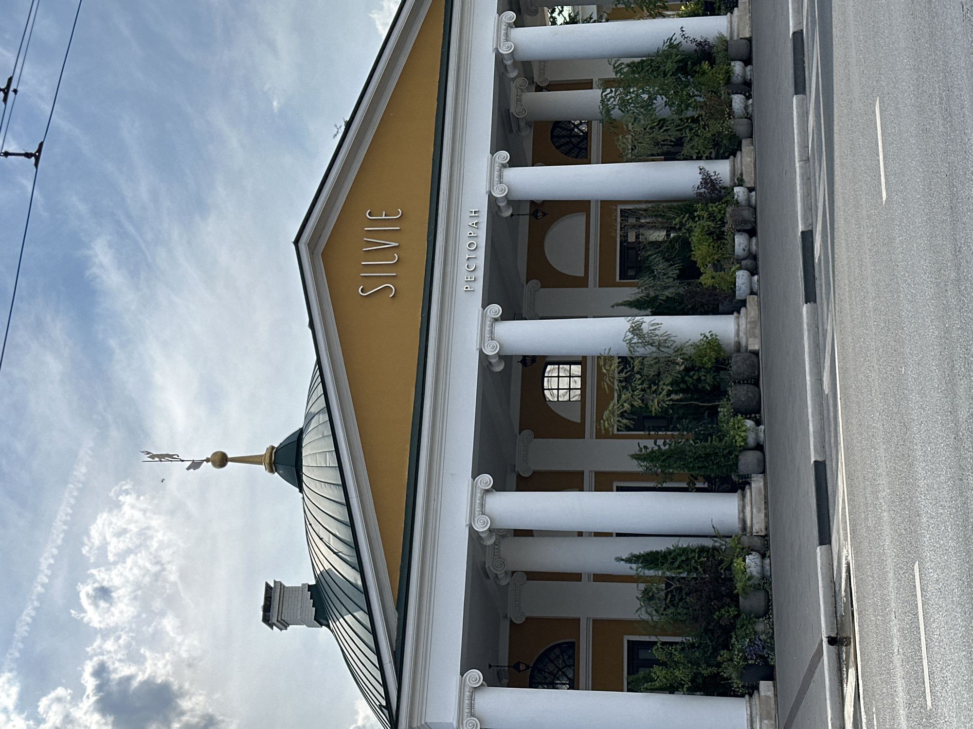 The image shows the facade of a restaurant called "SILVIE" in Golovinskoe, Russia. The building features a distinctive green domed roof with a golden weather vane topped by a figure. A white brick chimney is visible on the left side of the roof. The main entrance is adorned with a yellow facade, white columns with Corinthian capitals, and a prominent sign that reads "SILVIE" in large, metallic letters, with "РЕСТОРАН" (Restaurant) written below it in smaller letters. The building is surrounded by potted plants, which are arranged along the colonnade. A street with a white line marking the lane is visible in the foreground, with a sidewalk and curb separating it from the building. The sky above is partly cloudy with some wispy clouds and contrails. There are no people visible in the image.