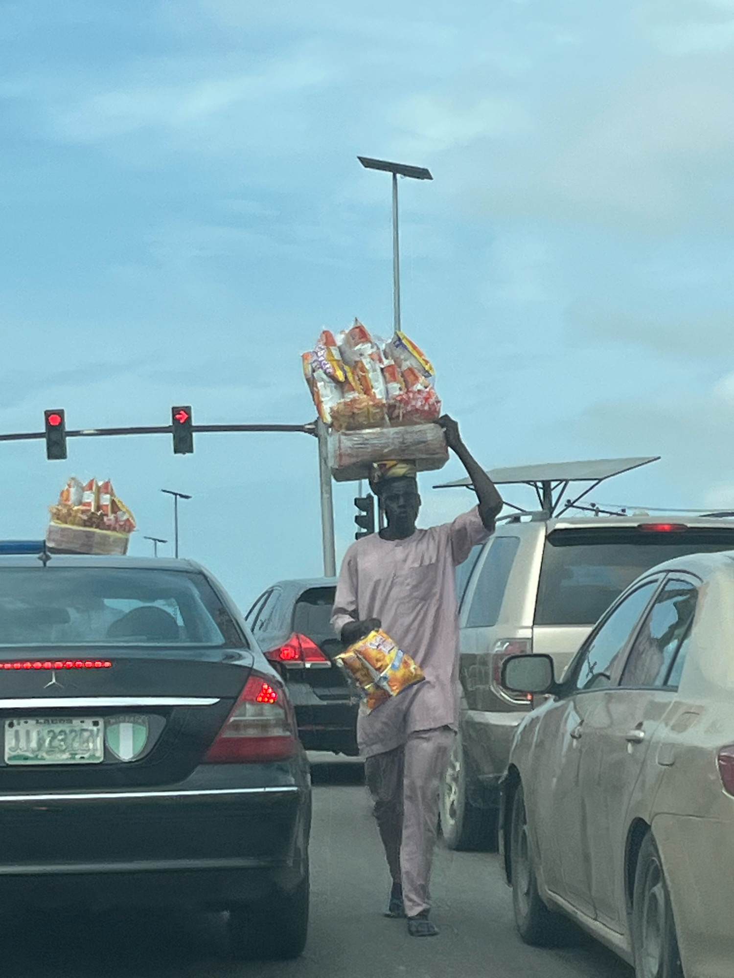 A man wearing a light pink tunic and pants balances a large stack of bagged snacks on his head while walking in traffic on the Lekki Peninsula II, Nigeria. He is walking alongside a dark Mercedes-Benz sedan and a light-colored SUV. A traffic light with a red signal and a red arrow is visible in the background. The sky is overcast with blue and gray clouds.