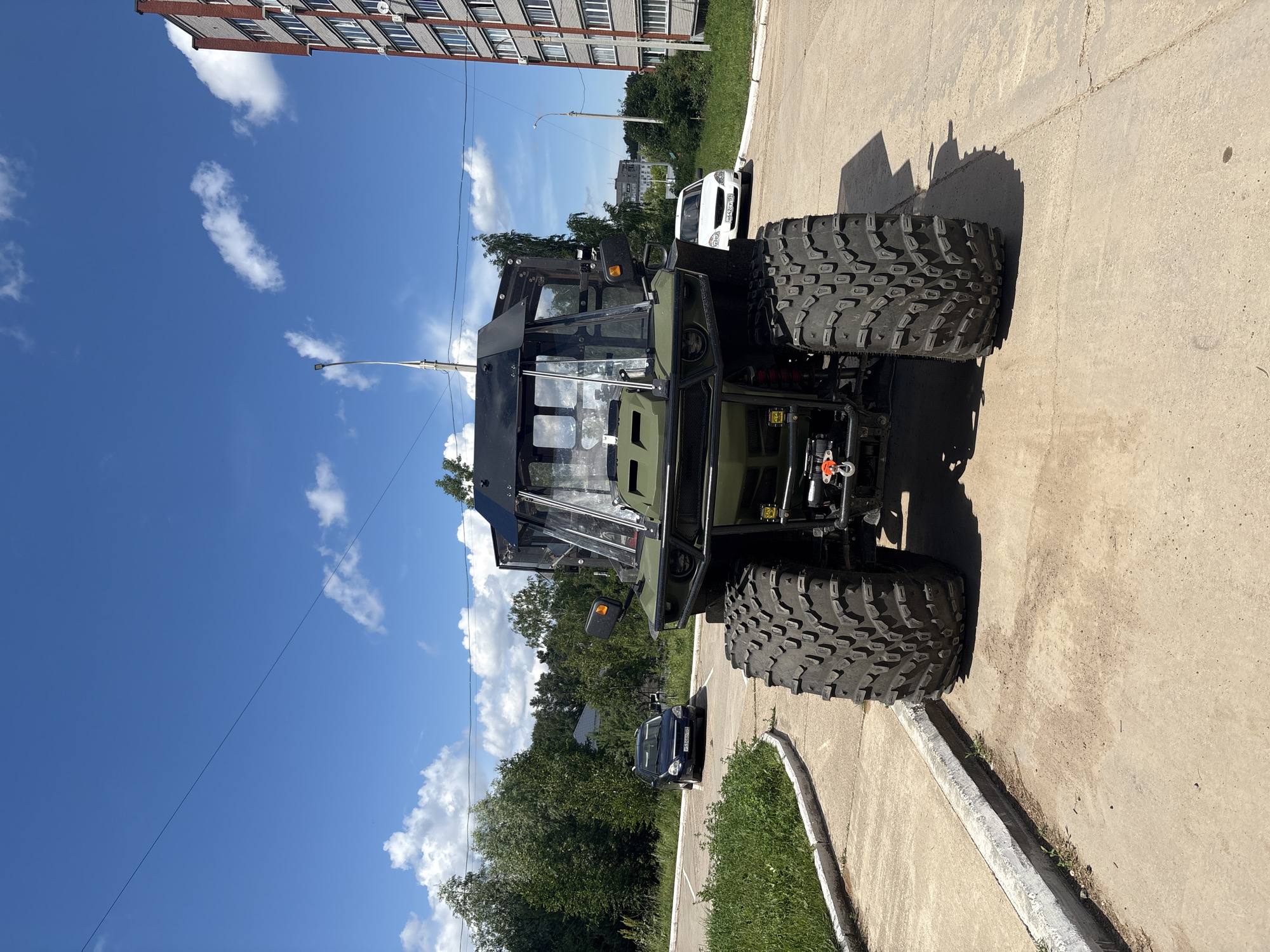 An all-terrain vehicle with large tires is parked on a concrete surface. The vehicle is dark green with a black cab and large windows. In the background, there is a residential building with several stories and a brick facade. Trees and a blue sky with white clouds are also visible. A dark blue car is parked in the distance to the left, and a white car is parked to the right. The scene appears to be in an urban or suburban area. The image was taken in Chaikovsky, Russia.