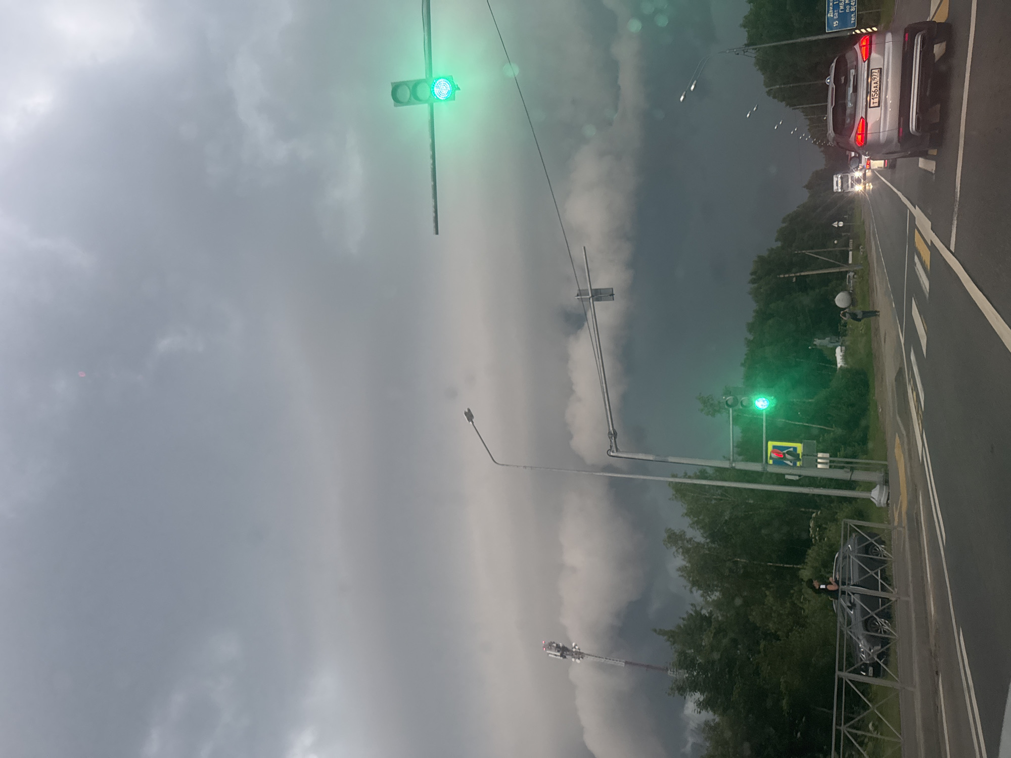 The image captures a street scene with a dark, cloudy sky, suggesting overcast weather. A traffic light is illuminated with a green signal, indicating traffic flow is permitted. In the foreground, a person is partially visible standing near a parked dark-colored car. Further down the road, several cars are seen, with one silver SUV prominently displayed in the right lane, its taillights illuminated. A street sign with Russian text is also visible on the right side of the road. The scene appears to be in a suburban or roadside setting with trees lining the area. The lighting suggests it is either early morning or late afternoon, given the overcast conditions and the intensity of the car headlights. The overall mood is somewhat somber due to the dark sky.
Visible text:
- T156AA97Z (license plate on the silver SUV)
- Движк... (partially visible on the street sign)
- 15 авг... (partially visible on the street sign)
- тел. 8(495)... (partially visible on the street sign)