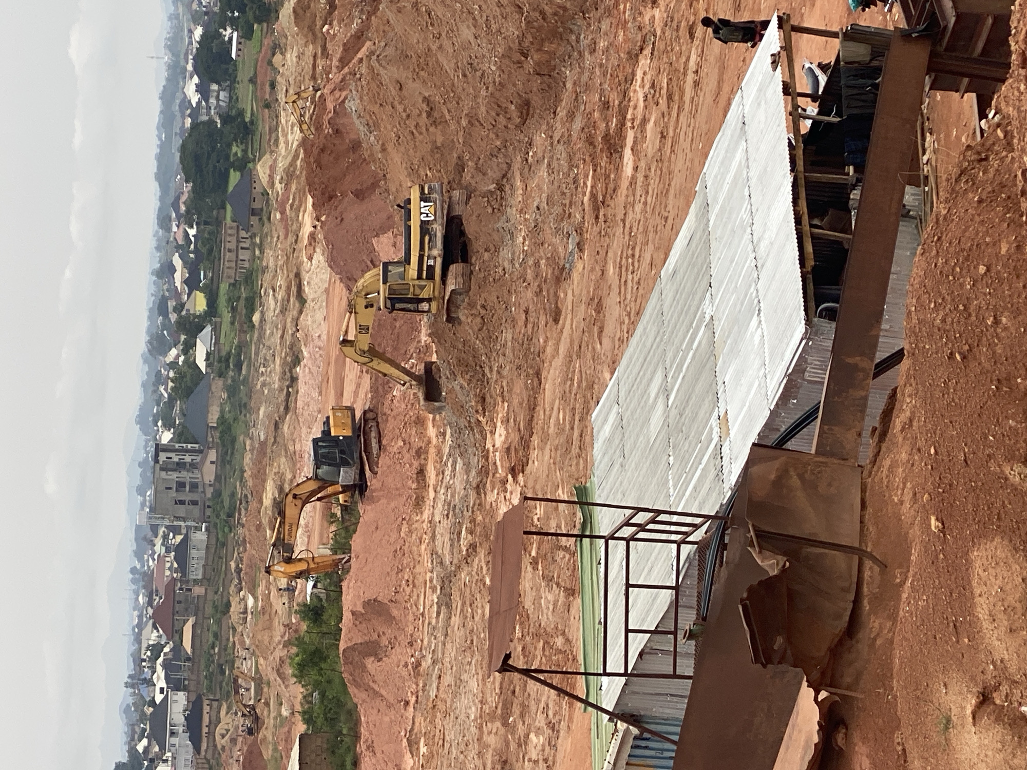 This is a mid-range shot of a construction site in Jos, Nigeria, with a landscape of red earth, excavated mounds, and a distant cityscape. In the foreground, there are several makeshift structures with corrugated metal roofs, one of which is partially collapsed and shows exposed wiring. Two large yellow excavators are actively working on the site, one in the middle ground and another further in the background. The excavators are yellow and black, with the "CAT" logo visible on one. There are a few individuals present: one person is standing near the corrugated structures, and another is walking away from them further up the hill. The cityscape in the background features numerous residential buildings with red and white roofs, interspersed with greenery and some taller structures. The sky is overcast, contributing to the muted lighting of the scene. The overall impression is one of ongoing construction and development in a semi-urban area.