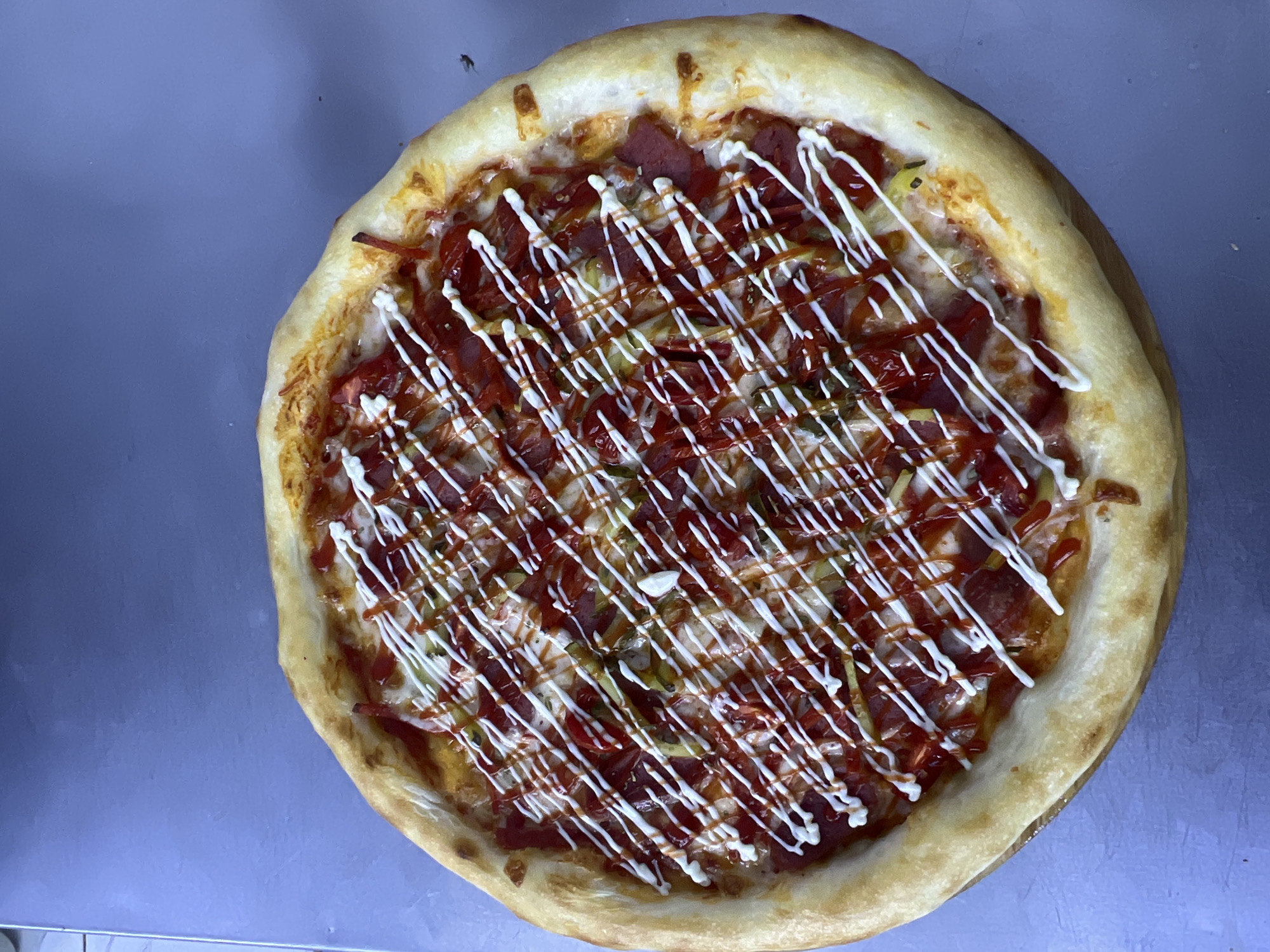 A top-down, eye-level shot shows a pizza on a gray surface. The pizza is round with a thick, golden-brown crust. The toppings include red sauce, white cheese, slices of what appear to be pepperoni or salami, and thin green slices that could be pickles or peppers. The pizza is finished with a drizzle of ketchup and a crisscross pattern of white sauce, possibly mayonnaise or ranch dressing. The background is a plain, matte gray surface. The scene appears to be indoors, likely in a kitchen or restaurant setting. There are no people or animals present in the image. There are no visible indications of the location being in Ақши, Kazakhstan.
