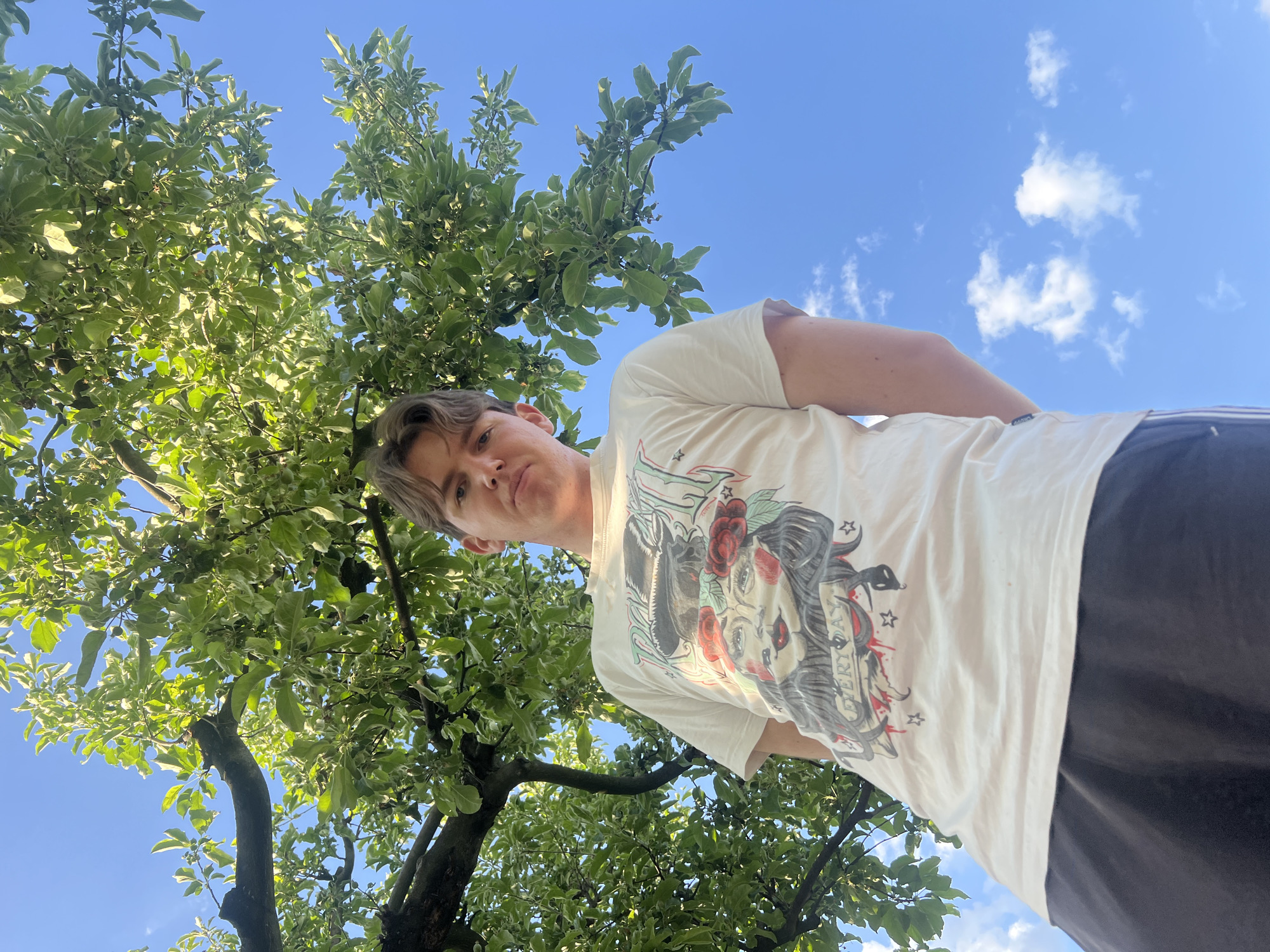 The image presents a low-angle view upwards towards a young adult male standing near a tree. The man has fair skin and light brown hair. He is wearing a light-colored t-shirt with a tattoo-style design on it. The tree, with green and light green leaves, occupies the left side of the frame. Above and around the tree and the man, there is a blue sky with sparse white clouds. The man appears to be looking down at the camera. Given the prompt, the location is likely Busmantsi, Bulgaria.