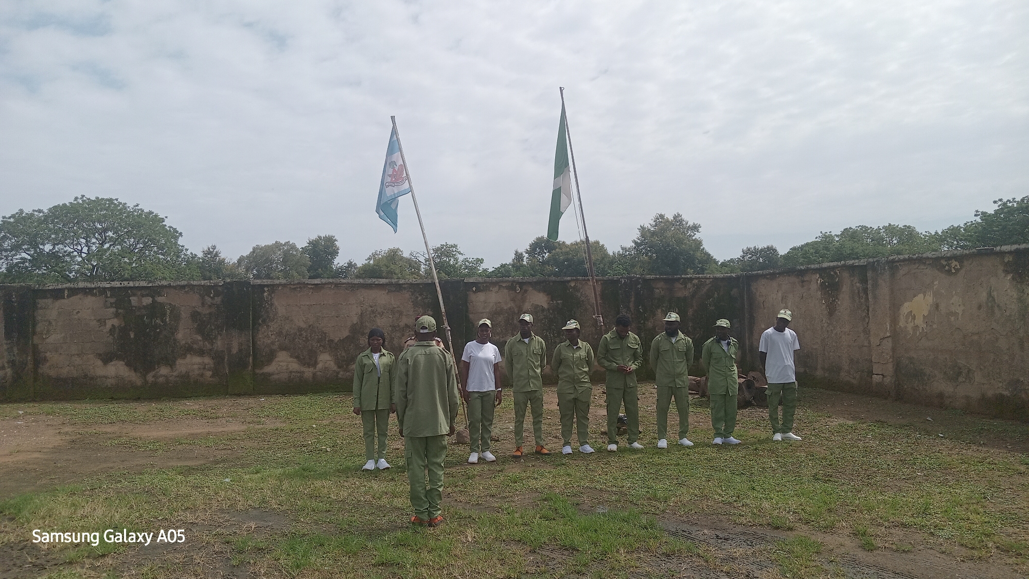The image captures a group of approximately nine individuals, most of whom are dressed in matching green uniforms and caps, standing in an open grassy area in front of a weathered, grey stone wall. Two flags are prominently displayed on flagpoles to the left of the group: one with a light blue and white design featuring a crest, and another larger flag which appears to be the Nigerian flag, with green and white stripes. The individuals are arranged in a line or formation, facing towards the left of the frame, with one person in the foreground facing away from the camera. The background consists of lush green trees and a cloudy sky, suggesting an outdoor setting. The overall atmosphere appears to be that of a formal gathering or ceremony. Based on the provided context, this scene is located in Toro, Nigeria.