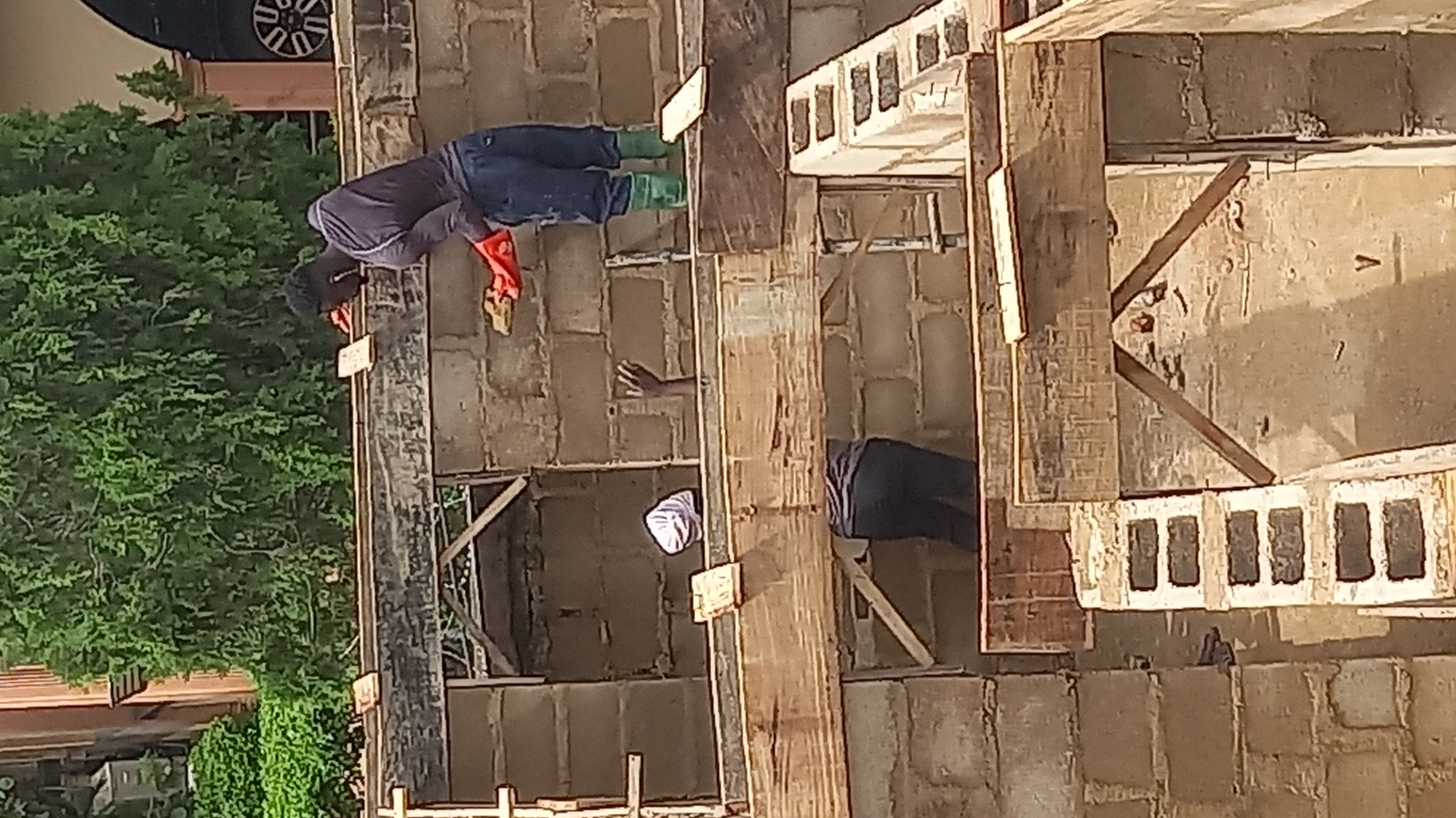 A construction site is depicted, possibly in Gura Top, Nigeria, featuring two individuals engaged in building activities. One person, wearing a dark long-sleeved shirt, blue pants, and green boots, is standing and holding a trowel with orange gloves. This individual appears to be applying mortar or cement to a wooden beam. Another person is partially visible within the structure, wearing a white head covering and dark pants. They are positioned lower than the first individual and are not clearly engaged in a specific action. The environment consists of concrete block walls and wooden scaffolding, indicating ongoing construction work. Lush green foliage is visible in the background, suggesting an outdoor setting.