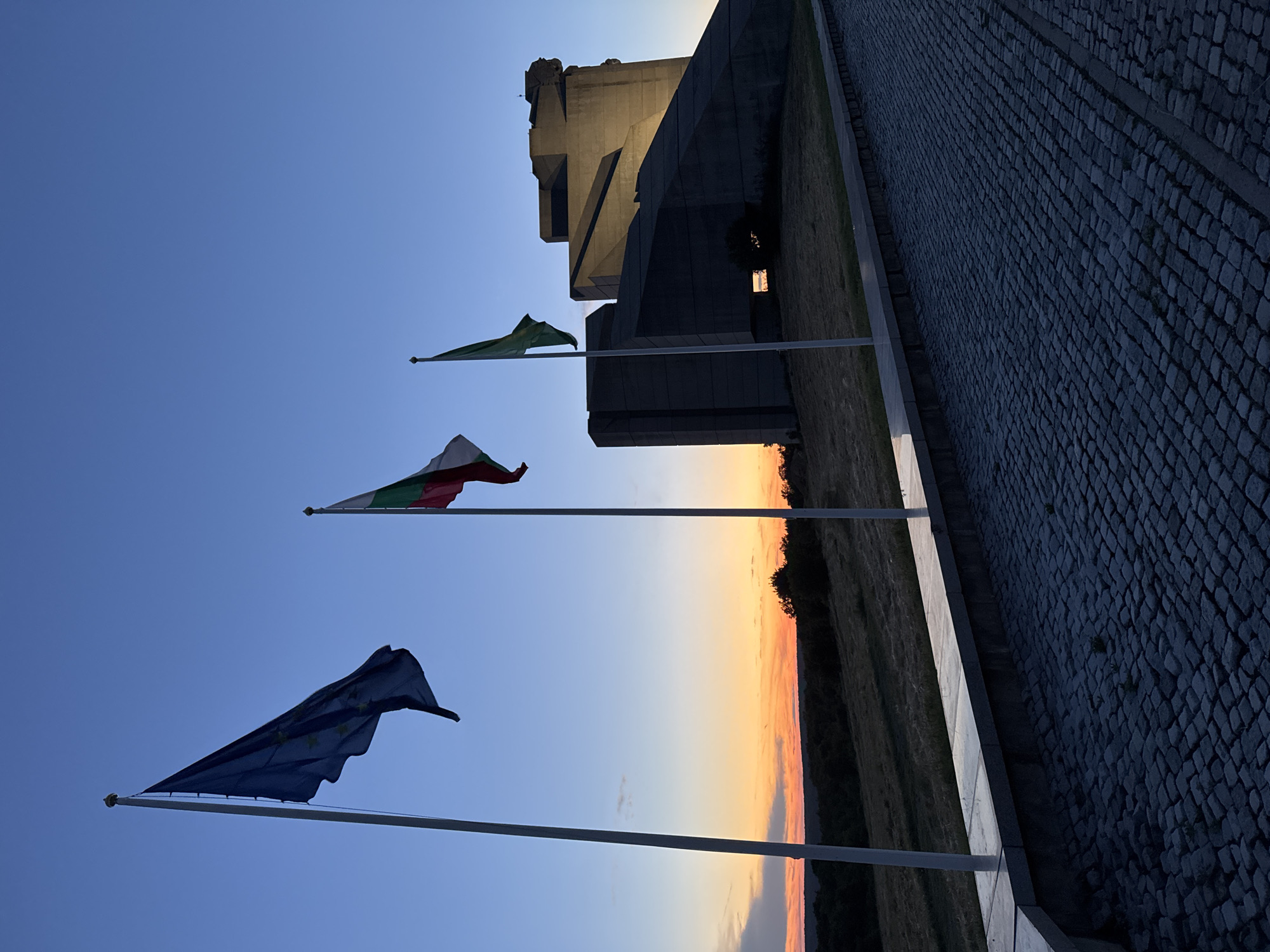 The image depicts the Monument to the Declared Founders of the Bulgarian State in Shumen, Bulgaria, at dusk. Three flags are prominently displayed on flagpoles in the foreground. The leftmost flag is the blue flag of the European Union, with its characteristic circle of yellow stars. To its right is the Bulgarian national flag, featuring three horizontal stripes of white, green, and red. The third flag, to the right of the Bulgarian flag, is green.
The monumental structure of the monument, constructed from concrete, is visible in the background. Its angular and imposing design is silhouetted against the twilight sky. The sky itself displays a gradient of colors, transitioning from a soft orange and yellow near the horizon, indicative of a sunset, to a deep blue overhead. The ground in the foreground is paved with dark cobblestones, creating a textured pathway. A low, stone wall with a granite top borders the cobblestone area. Beyond the flags and monument, a grassy slope stretches towards a distant, dark tree line, also silhouetted against the colorful sky. The overall scene conveys a sense of solemnity and national pride under the fading light of day. There are no people visible in the image.
