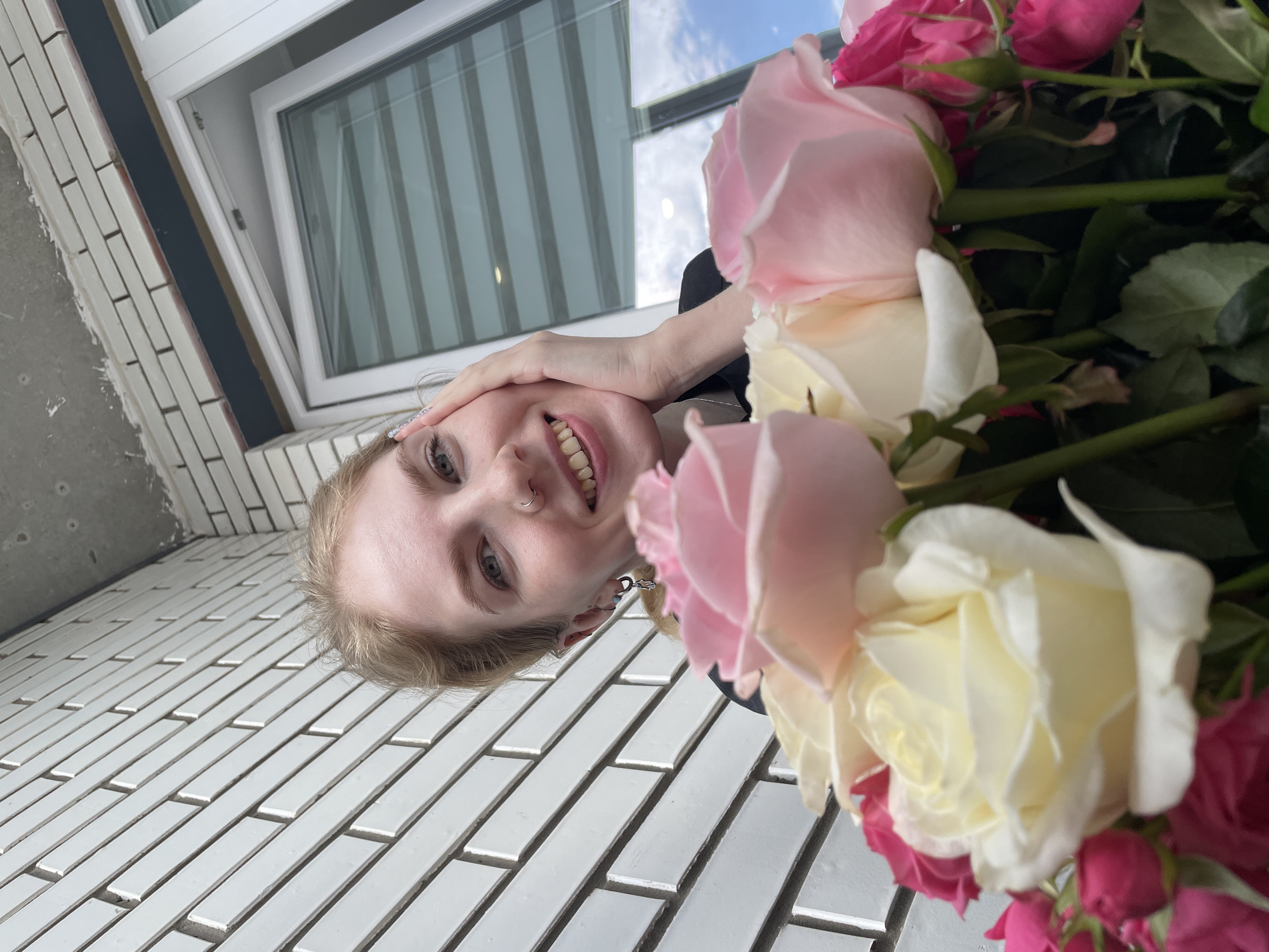 A young woman is captured in a vibrant, tilted portrait, smiling broadly with an expression of joy. Her blonde hair frames her face, revealing light eyes and a nose piercing. She has her left hand raised, gently touching her forehead.
The foreground and right side of the image are dominated by a lush bouquet of roses, featuring blossoms in rich pink, pale pink, and creamy white hues. Their fresh petals and green stems add a touch of natural beauty to the scene.
The background consists of a surface covered with small, rectangular white tiles arranged in an offset pattern, possibly a wall or floor. Above and behind the woman, a modern white-framed window or skylight is visible, through which a bright, cloudy daytime sky can be seen. The unique low-angle and tilted perspective give the photograph a dynamic feel. The overall impression is one of happiness and celebration, with no visible text in the image. The location is specified as Izhevsk, Russia.