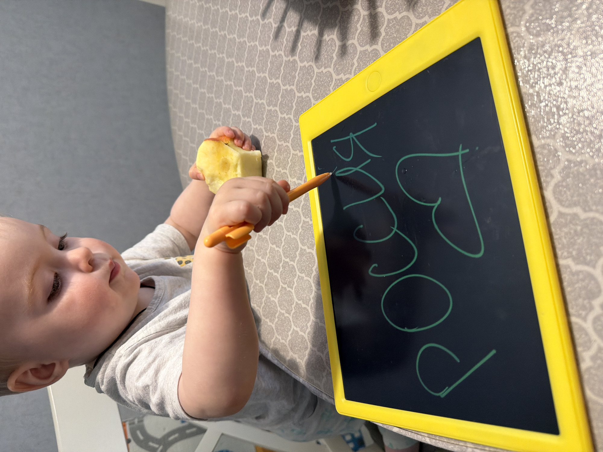 A toddler is positioned beside a yellow-framed electronic drawing tablet, held at an angle. The child is fair-skinned, wearing a light grey shirt, and holding a partially eaten apple in their left hand. Their right hand holds an orange stylus, which they are using to write on the tablet. The tablet displays green markings that appear to spell out "poco". The table beneath has a patterned grey tablecloth. The scene appears to be indoors. The location is not discernible as Votkinsk, Russia, from the given image.