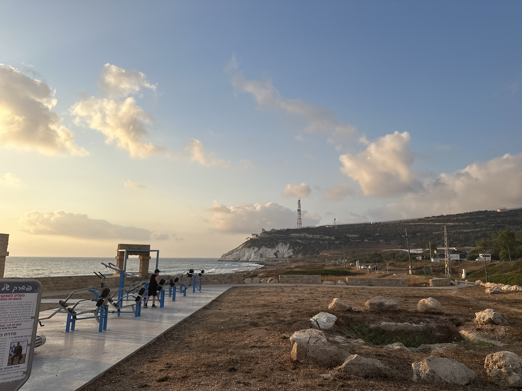 The image depicts an outdoor exercise area situated along the coast of the city of Mate Asher Regional Council, Israel. In the foreground, a row of blue and metal exercise machines is arranged on a paved walkway. A single person, dressed in black athletic wear, is positioned on one of the machines, appearing to be mid-workout. The sea is visible in the background, with gentle waves lapping against the shore. Beyond the coastline, a rocky cliff face rises, topped with vegetation and a communication tower. The sky is a blend of blue and white, with scattered clouds catching the warm light of what appears to be either sunrise or sunset. The ground around the exercise area is covered in dry grass and scattered rocks. A sign with Hebrew text is present on the left side of the frame.