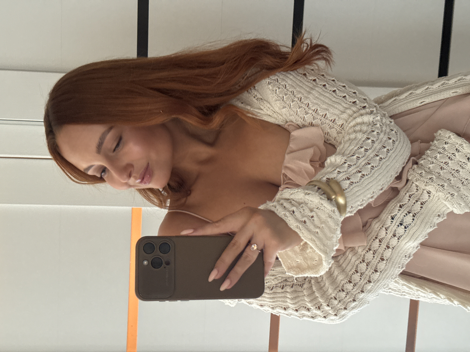 A medium shot shows a woman with auburn hair taking a selfie in a well-lit room with white and light gray walls. She is wearing a pale pink, ruffled dress and an off-white, crocheted cardigan. On her left hand, she wears a silver ring with a light-colored stone. She is holding a dark brown smartphone in her right hand, angled to capture her reflection. A wide, gold bracelet adorns her left wrist. The background features vertical white and black stripes on the right side of the image and a horizontal orange stripe on the left. There is no indication of the specific location in the city of Chaikovsky, Russia.