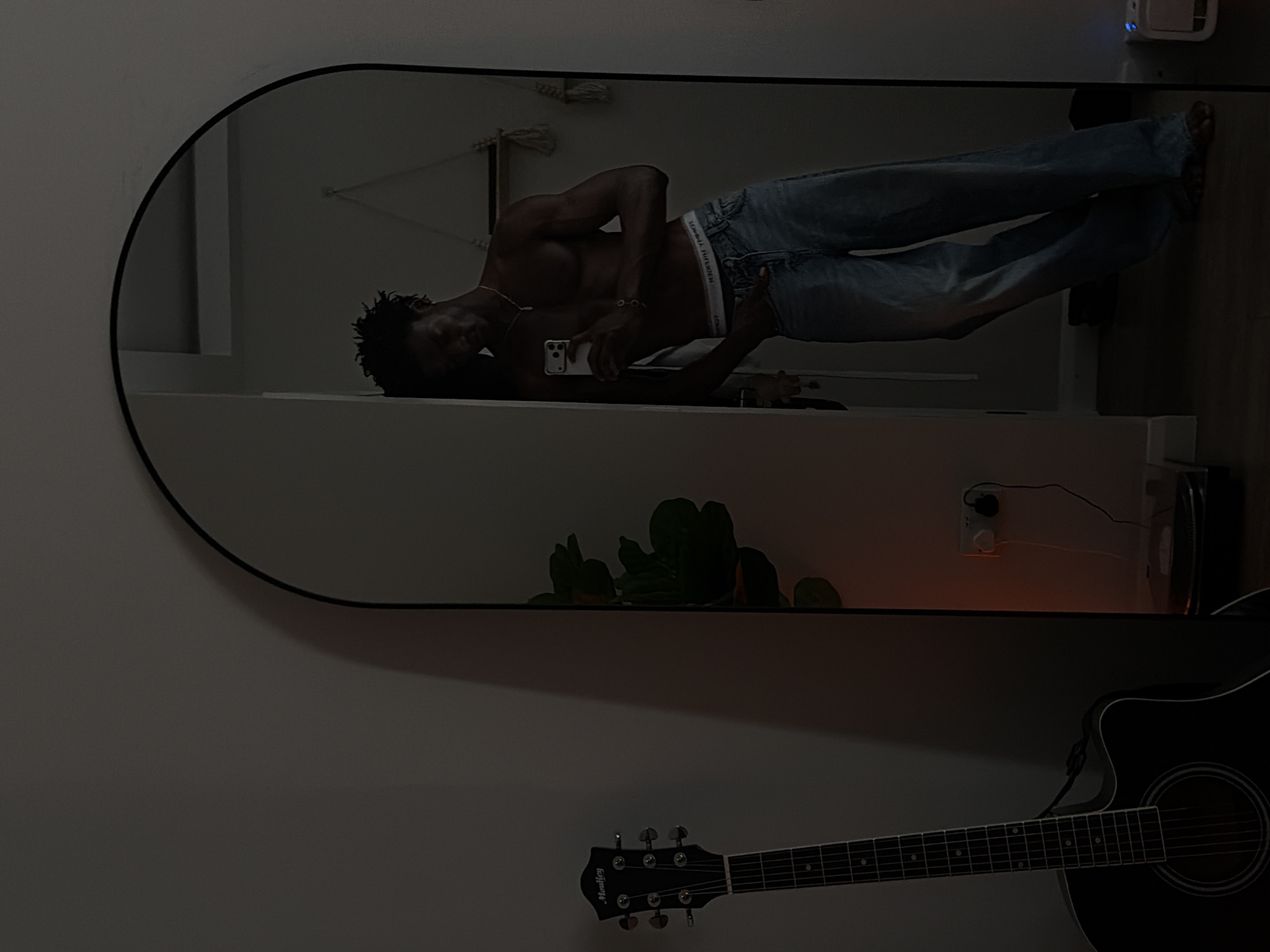 A young Black man is reflected in a tall, arched mirror, leaning against the frame. He is shirtless, wearing blue jeans and white underwear. He holds a phone in his right hand, appearing to take a selfie. His left hand is in his front pocket.
To the left of the mirror, an acoustic guitar with a black body and neck stands upright against a plain wall. A large houseplant with dark green leaves is visible in the reflection, positioned behind the guitar. In the background of the reflection, a white wall with a small wooden shelf is visible, adorned with two macrame hangers. There is an electrical outlet with two plugs connected to devices on the wall near the floor.
The overall lighting suggests it is evening or nighttime, with dim ambient light creating shadows. The setting appears to be an indoor domestic environment, possibly a bedroom or living area. There is no information within the image to suggest the location is in London, United Kingdom.