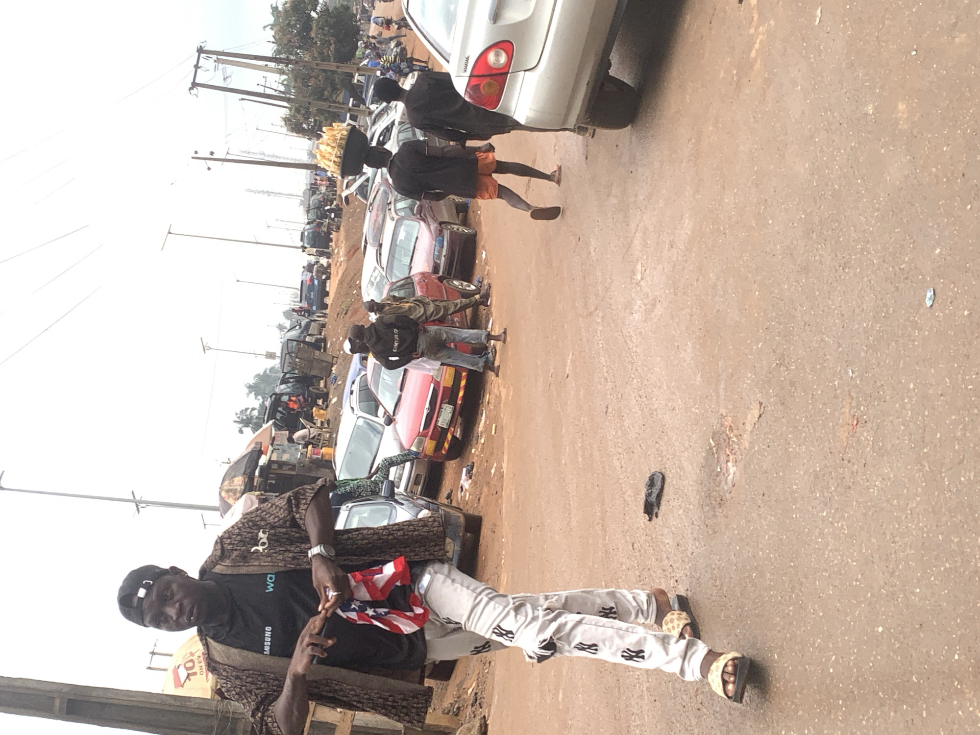 The image depicts a street scene in Shagamu, Nigeria, likely a market or roadside area with parked vehicles and a few individuals. In the foreground, a man wearing a black cap, a patterned jacket over a black t-shirt, and white pants with black "NY" logos walks towards the viewer. He holds a small American flag in his right hand and a cigarette in his mouth. His left hand is visible, holding what appears to be a small object.
Behind him, several cars are parked on a dirt road. To the right, two men are walking away from the camera. One is wearing a black t-shirt and orange shorts, carrying a large basket of food on his head. The other is wearing a black t-shirt and black shorts. A silver car is prominently visible in the right foreground, partially obscuring another vehicle. In the background, more parked vehicles and market stalls can be observed, along with power lines and utility poles. The overall impression is that of a busy, informal commercial area.