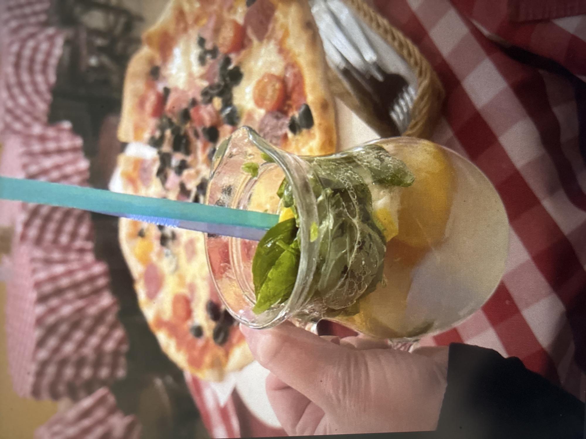 An overhead shot captures a close-up of a hand holding a glass pitcher filled with a refreshing drink. The pitcher contains ice, slices of lemon or orange, and fresh mint leaves, with a vibrant blue straw emerging from the top. In the background, a partially visible pizza sits on a white plate, adorned with pepperoni, olives, and cherry tomatoes. The scene is set against a red and white checkered tablecloth, hinting at a casual dining atmosphere. The context of Novosibirsk, Russia, is not discernible from the visual elements of the image.