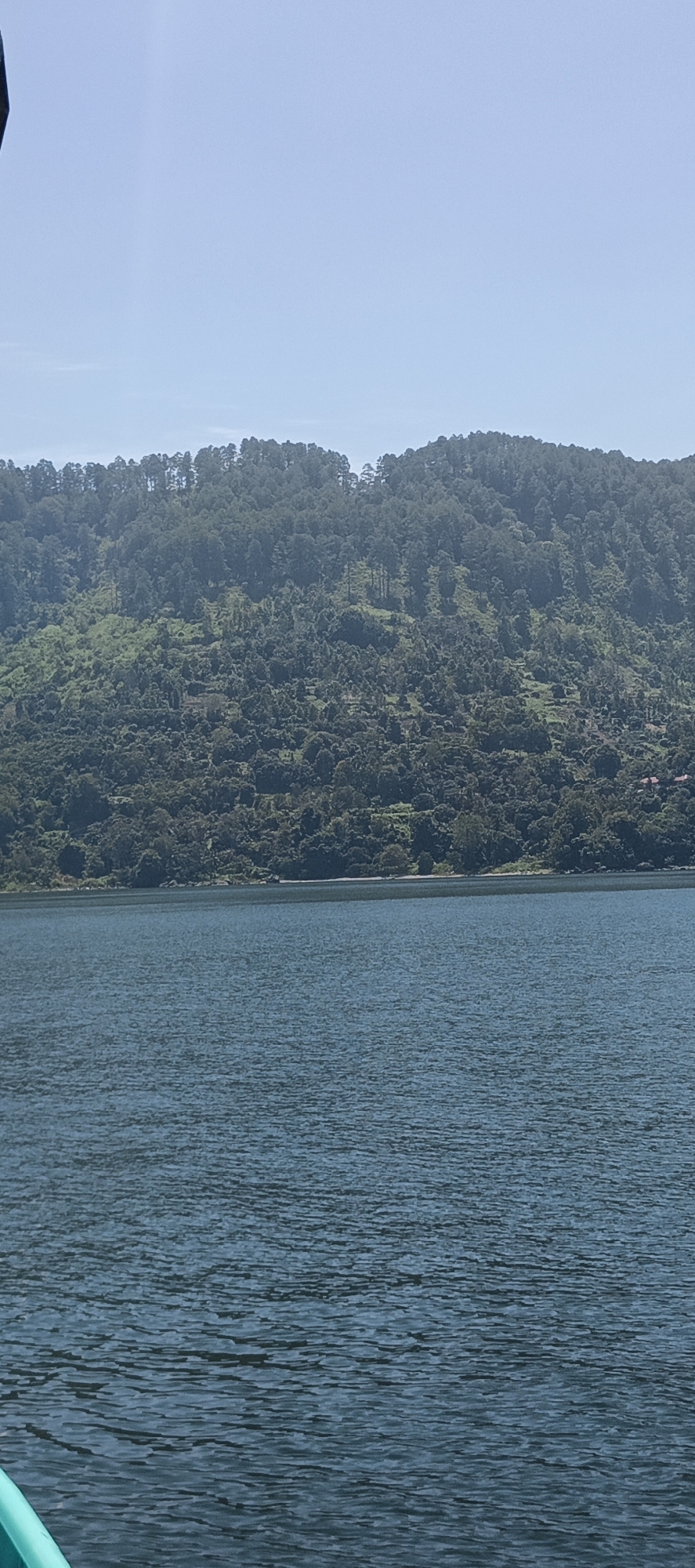 The image shows a body of water in the foreground, with a densely forested hillside rising in the background. The sky is clear and pale blue. The water is a deep blue-green, with ripples on the surface. The hillside is covered in a variety of trees, with some patches of lighter green that may indicate vegetation or clearings. The overall impression is one of a natural, somewhat remote landscape. The location is the city of Tigaras, Indonesia.