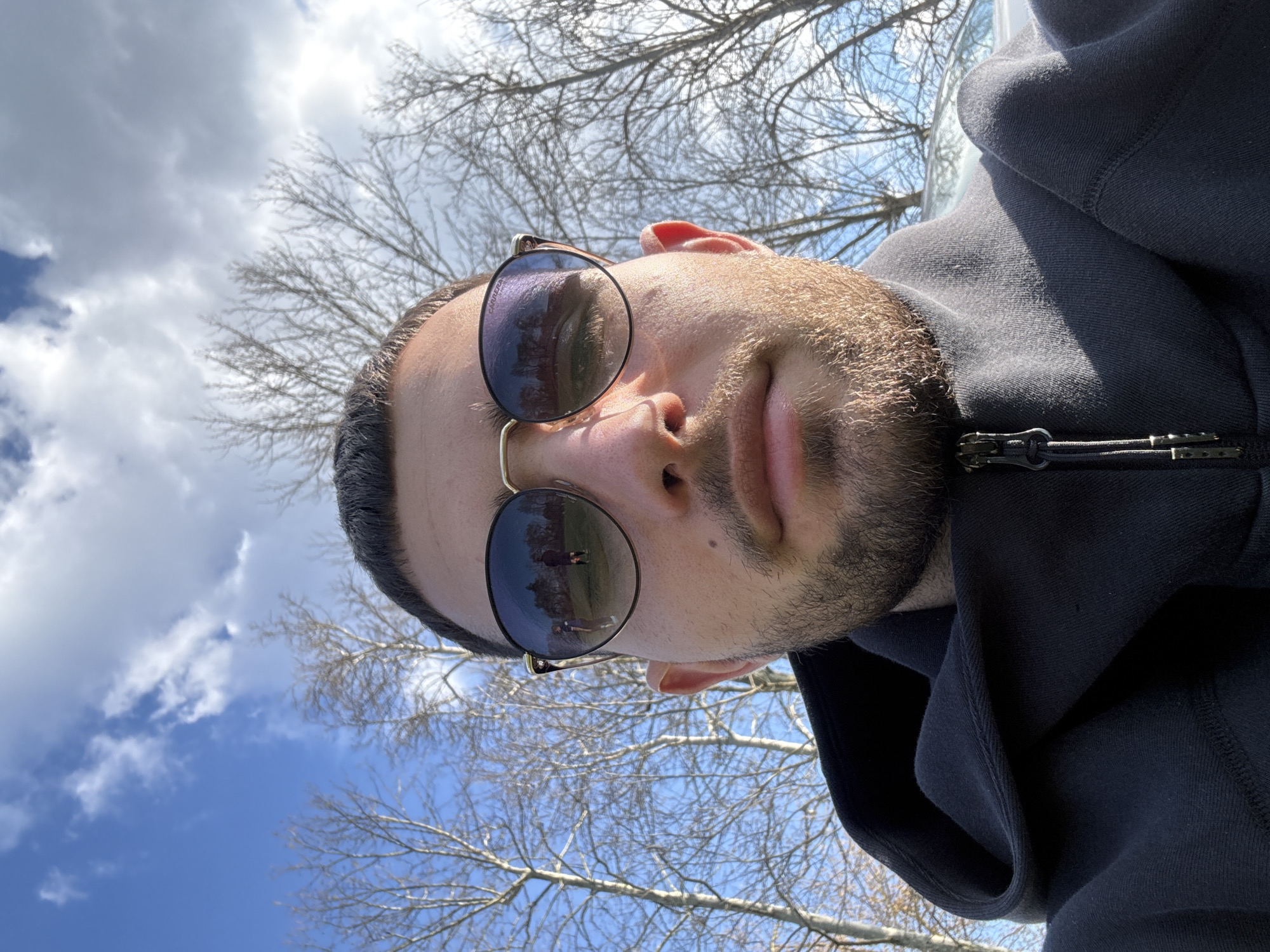 A man is the subject of the photograph. He is wearing sunglasses, and a black hooded sweatshirt. The background features a blue sky with white clouds and bare tree branches. The reflection in his sunglasses contains people standing on a field of green. The exact location in the city of Dushevo, Bulgaria is indeterminable from the image alone.