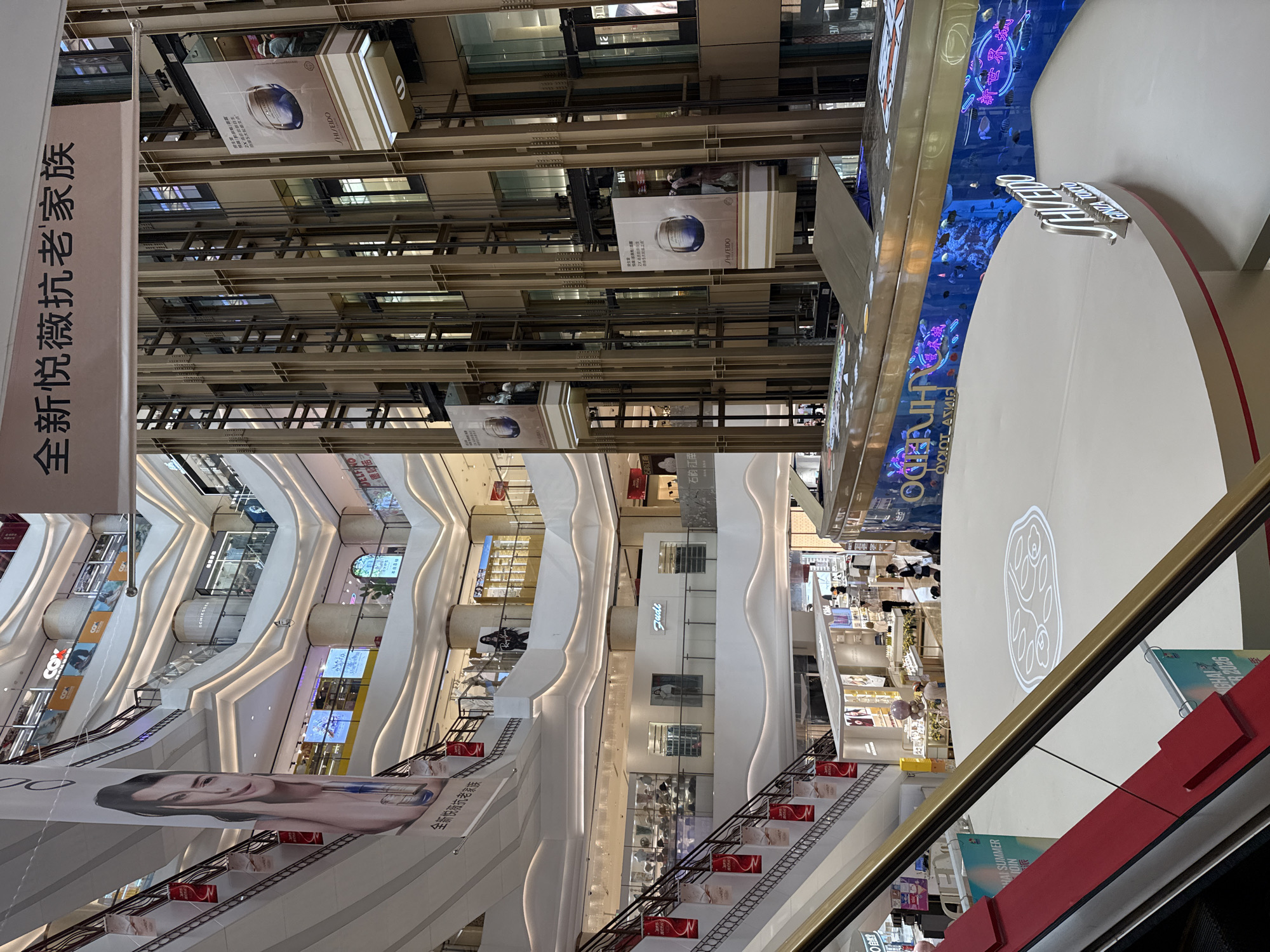 This image captures a bustling multi-level interior of a modern shopping mall in Shanghai, China. A large, multi-story atrium dominates the scene, with escalators and open walkways connecting various floors. The architecture features sleek, curved white surfaces accented by warm lighting, creating a sense of spaciousness and luxury.
Several advertising banners are prominently displayed. One large banner on the left features a close-up of a woman's face and text in Chinese characters. On the right, suspended banners advertise Shiseido, a skincare brand, with images of their products. A glass-enclosed elevator shaft with metal framing is visible in the center of the atrium, with elevator cars moving between floors.
The mall appears to be active, with several individuals visible on different levels, browsing shops, or walking. Some shops have glass storefronts displaying merchandise. In the foreground, a large, circular white platform is set up, likely for an event or display, with a projection of a logo on its surface. Adjacent to this platform is a long, curved structure with an aquarium visible inside, filled with colorful fish and illuminated by neon lights. The text "SHISEIDO GINZA TOKYO" is prominently displayed on this structure.
The overall impression is one of a vibrant and upscale retail environment, characteristic of a major commercial hub in Shanghai.