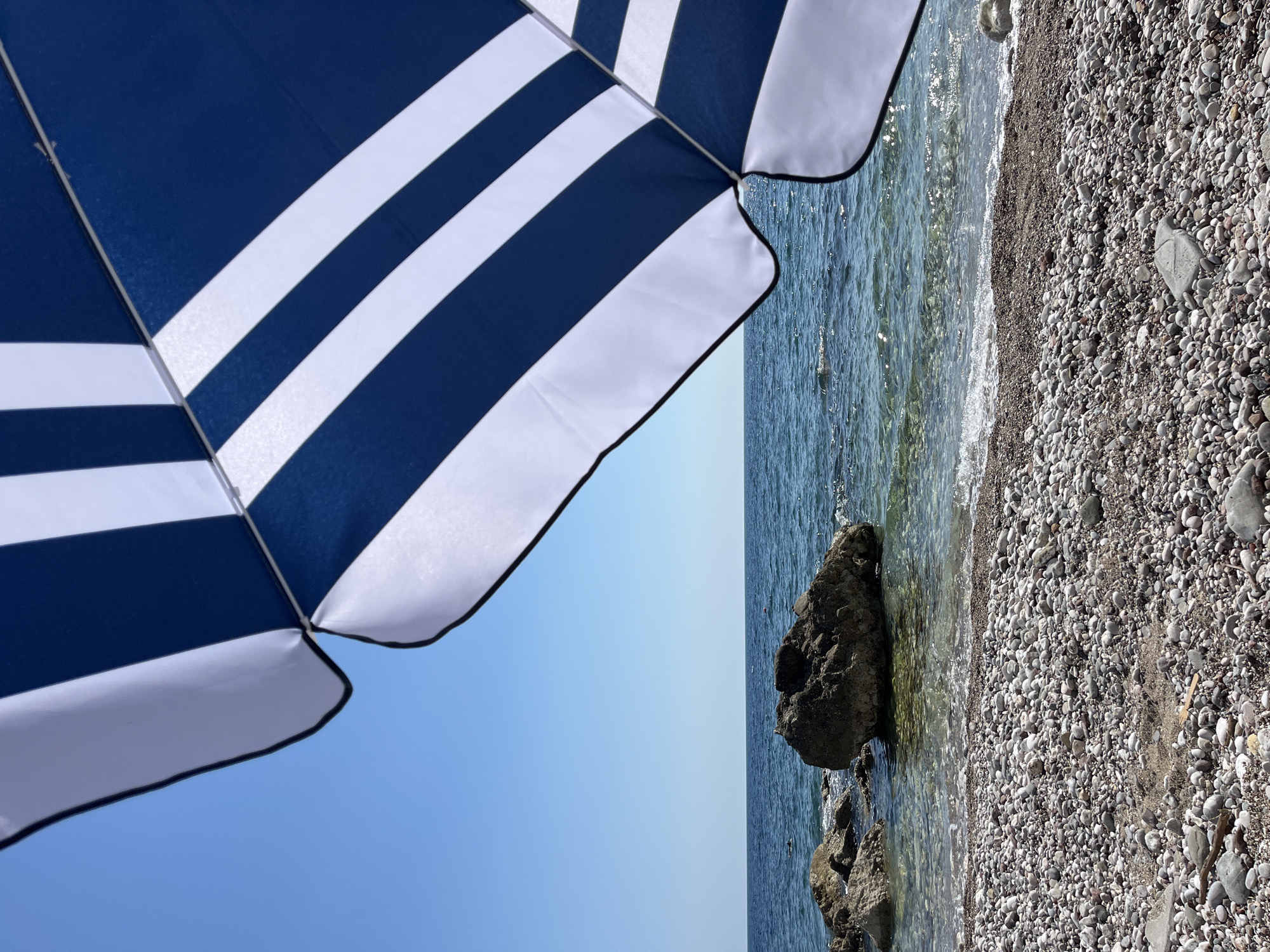 In the city of Bar, Montenegro, a beach umbrella with blue and white stripes is positioned in the foreground, casting a shadow over a pebbly shore. The calm, clear blue water of the Adriatic Sea stretches to the horizon under a cloudless sky. Several large rocks are visible partially submerged in the shallow water near the shore, and a few scattered individuals can be seen swimming or floating in the distance. The overall scene depicts a peaceful day at the beach.