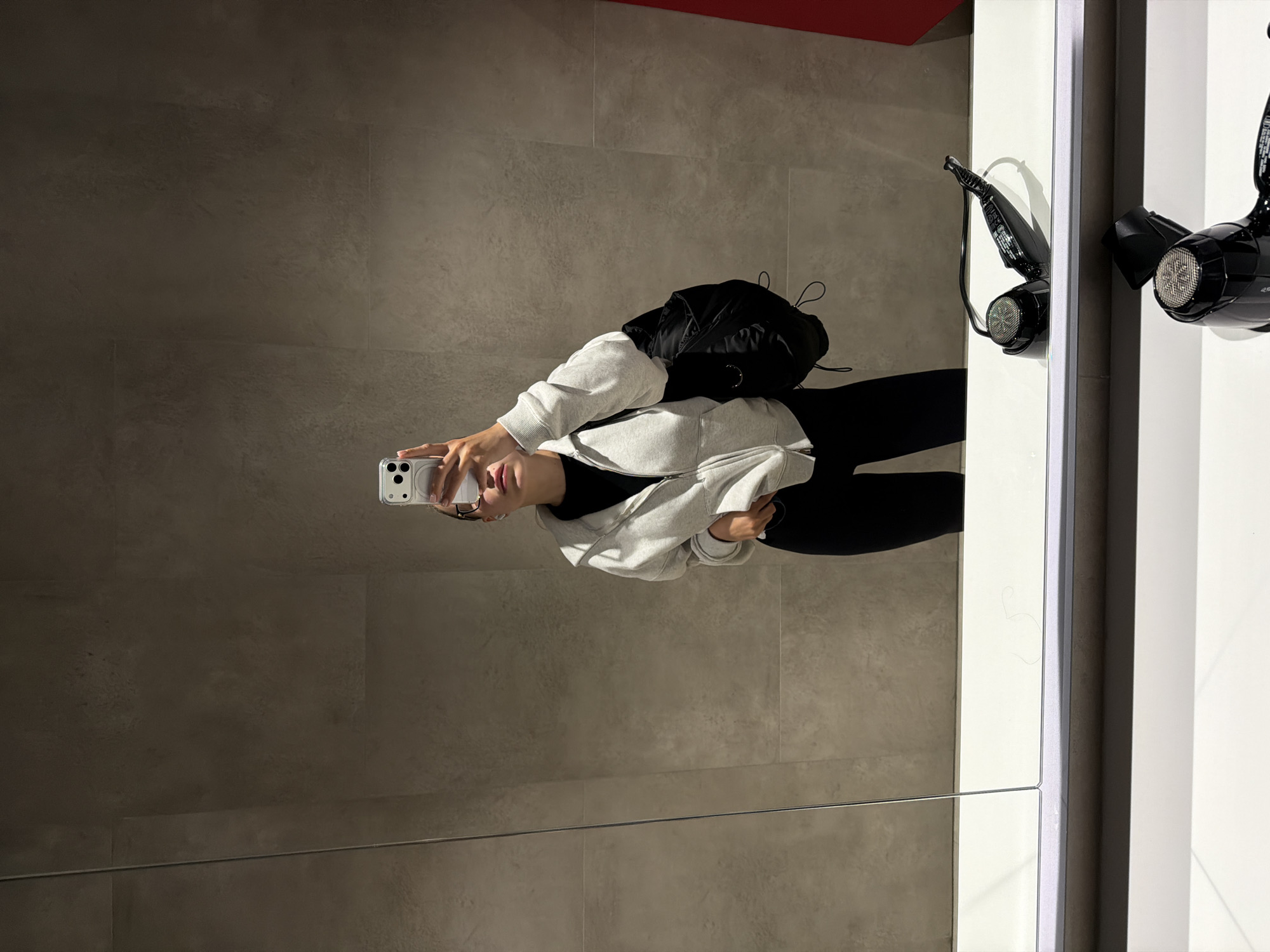 A mirror selfie in a bathroom-like space shows a person holding a smartphone to take a picture. The individual is dressed in a light gray hoodie and black leggings with a black backpack. The background features gray tile walls. To the right of the reflection, two black hair dryers are mounted on a white wall. A red element is seen at the top. The location in Podolsk, Russia is not known.