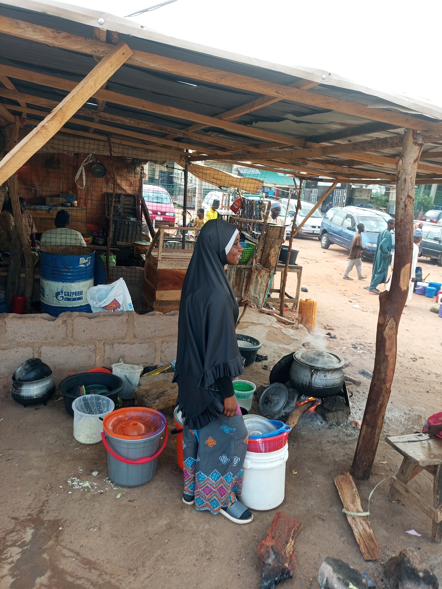 In the city of Gombe, Nigeria, a street food vendor, a woman in a black hijab and a patterned skirt, is seen tending to a cooking fire under a makeshift shelter. Large pots are placed on the fire, with steam rising from one of them, indicating active cooking. Various buckets, basins, and cooking utensils are scattered around the immediate area. The shelter is constructed with a wooden frame and a corrugated metal roof, supported by a rough-hewn tree trunk. In the background, a dirt road is visible with several vehicles, including cars and a blue minivan, suggesting this is a busy thoroughfare or marketplace. Other people are present in the background, some appearing to be customers or passersby. The overall scene depicts a typical street-side food preparation area in a bustling Nigerian urban environment.