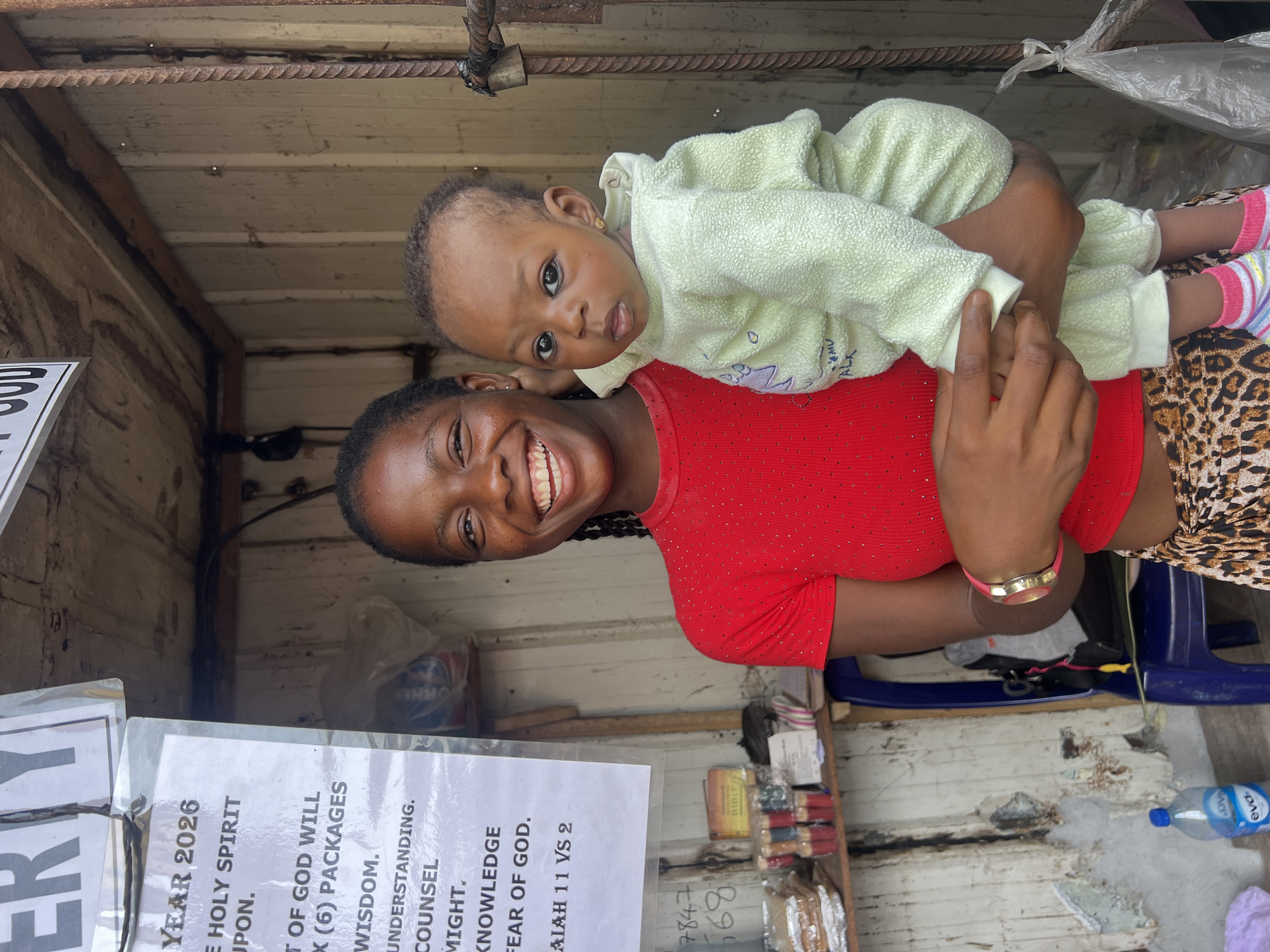 In Nkpor, Nigeria, a young woman with dark skin and braided hair, wearing a red, sparkly top and a leopard-print skirt, stands in a narrow stall. She is smiling and holding a baby dressed in a pale green, textured outfit. The baby has dark eyes and is looking towards the camera. Behind them, a white sign is attached to the stall's corrugated metal wall, listing items like "YEAR 2026," "HOLY SPIRIT," and biblical references such as "ISAIAH 11 VS 2." To the left, shelves display various items, including what appear to be small packages, candles, a Bible, and a bottle of Eva water. The stall's interior walls are made of weathered corrugated metal. A blue plastic chair is visible to the left. A padlock is attached to a rusty metal bar on the right side of the frame.