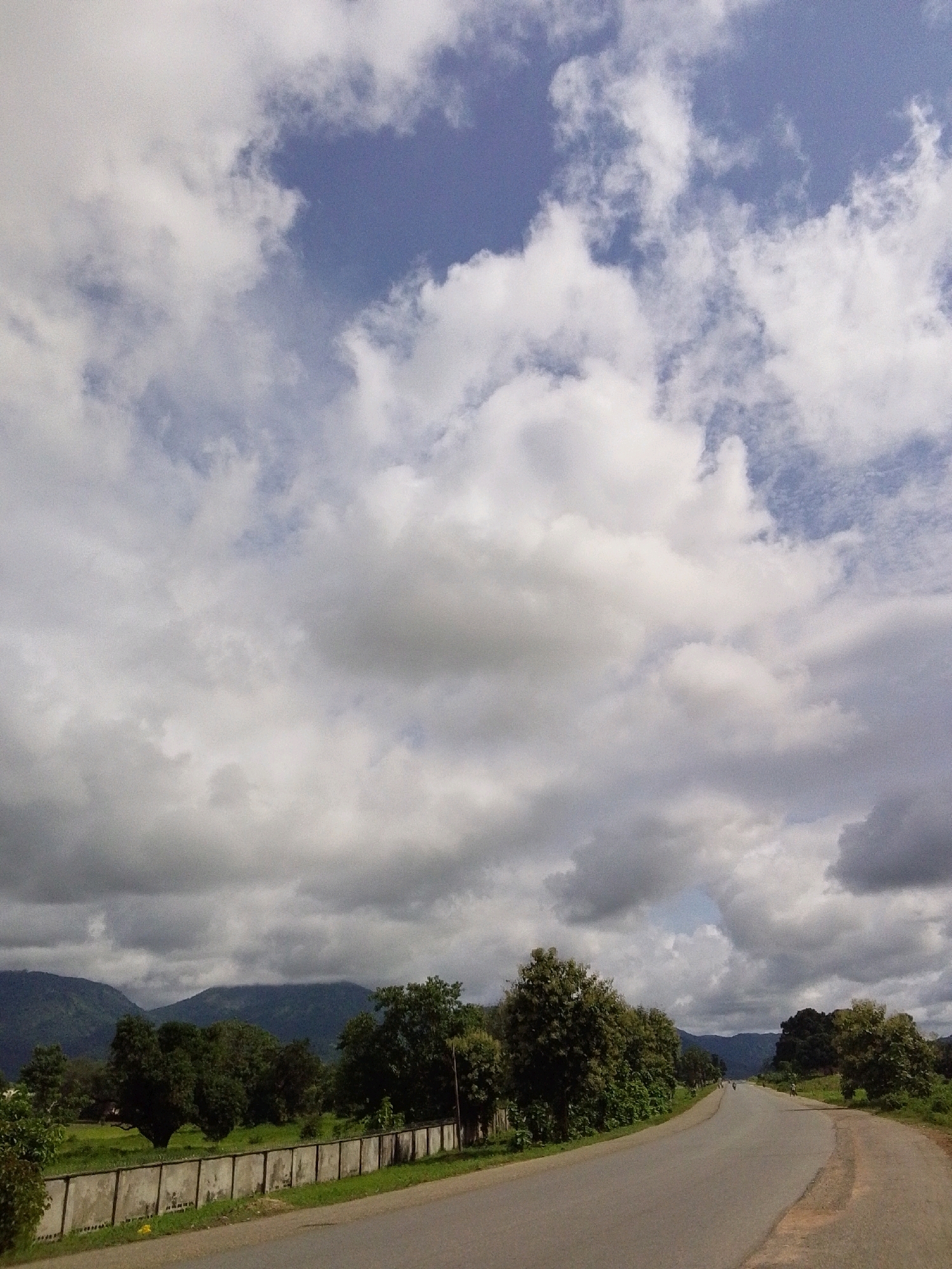 A road curves into the distance through a rural landscape. The sky is filled with dramatic, fluffy white and gray clouds. On either side of the road are lush green trees and vegetation. In the background, rolling hills covered in trees rise up. A low, concrete wall runs along the side of the road in the foreground. Two people on motorcycles are visible on the road in the distance.