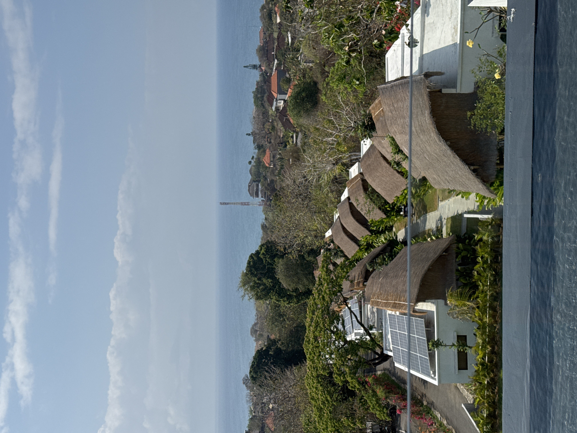 The image depicts an aerial view of a resort complex situated in Kuta Selatan, Indonesia. The foreground is dominated by a body of water, likely a swimming pool, with its shimmering surface reflecting the bright daylight. Beyond the water, a row of bungalows with thatched roofs is visible, nestled amidst lush green vegetation. Some of the bungalows have modern white structures attached, some of which are fitted with solar panels. A paved pathway winds through the resort, connecting the structures.
In the mid-ground, dense tropical trees and foliage create a vibrant green canopy, interspersed with some bare branches indicating a dry season or specific tree types. The landscape gently slopes downwards, revealing a scattered settlement with houses featuring red-tiled roofs and white walls. A tall communication tower stands out against the horizon, symbolizing the presence of infrastructure in the area.
The background is a vast expanse of the ocean, its blue waters meeting a hazy sky with scattered white clouds. The overall scene captures a serene tropical setting, characteristic of coastal resorts in Southeast Asia, with a blend of natural beauty and man-made structures. There are no people visible in the image.