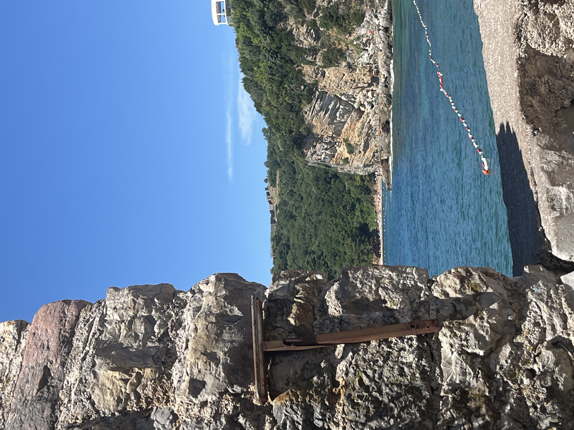 In Budva, Montenegro, a section of an old stone wall, partially covered in moss and featuring a rusty metal bracket, dominates the left foreground of the image. Beyond the wall, a bright blue sky stretches above a picturesque bay. The water is a clear, vibrant turquoise, marked by a line of red and white buoys. On the right side of the bay, a rocky, tree-covered cliff rises, with a white, modern structure visible at its summit. Below the cliff, a small beach with numerous people can be seen. Further in the distance, across the bay, more hilly terrain is covered in dense green vegetation, with ancient ruins visible on a distant hilltop. The overall scene depicts a sun-drenched coastal landscape.