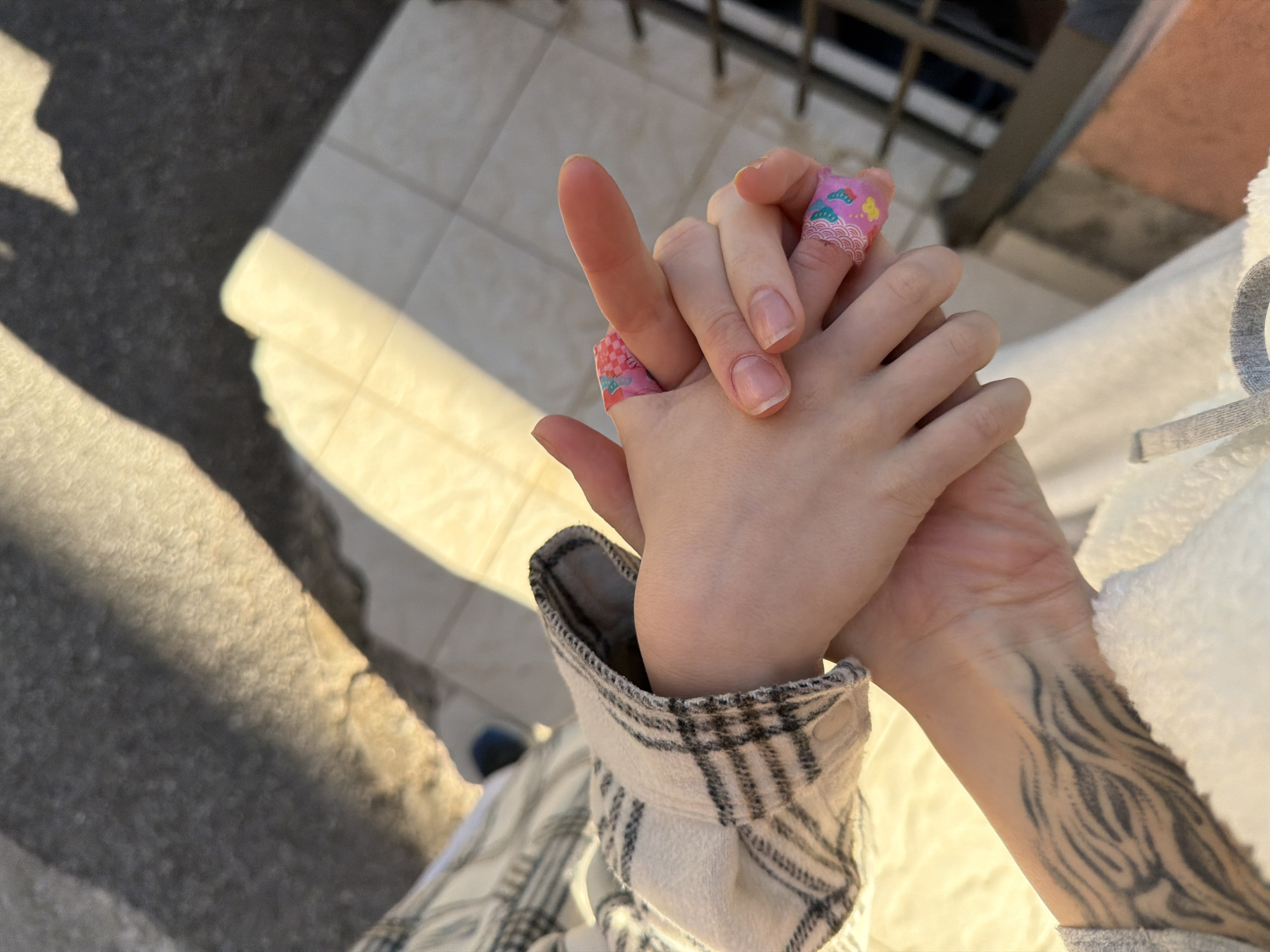 The image shows two hands clasped together. One hand is smaller and has two fingers wrapped in decorative bandages. The other hand is larger and has visible tattoos on the wrist and forearm. The person with the larger hand is wearing a plaid shirt and a white, fluffy jacket. The background is blurry but appears to be an outdoor setting with a tiled surface. There is no evidence in the image that it is related to the city of Medinski Krš, Montenegro.