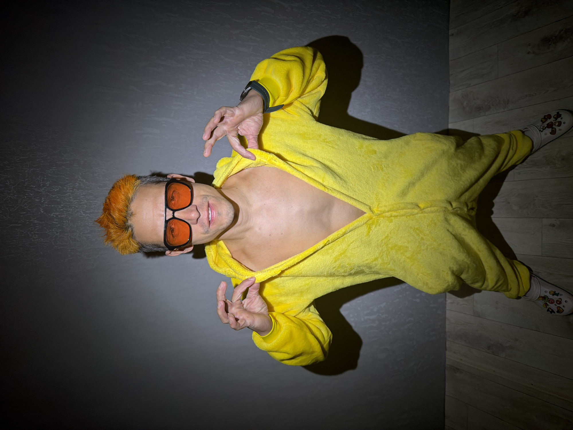 A medium shot captures a man dressed in a yellow, possibly plush, one-piece garment, similar to a jumpsuit or onesie. The garment is unzipped, revealing a portion of his bare chest. He sports orange-tinted hair and sunglasses with orange lenses. He makes a gesture with both hands, forming loose "okay" signs. He's wearing a dark-colored watch on his left wrist and white socks with decorative shoes, possibly Crocs. The background consists of two planes: a dark grey wall and a lighter wooden floor. The image is oriented at an angle, and shadows suggest a single light source. No information about the location of Chaikovsky, Russia is discernible in the image.