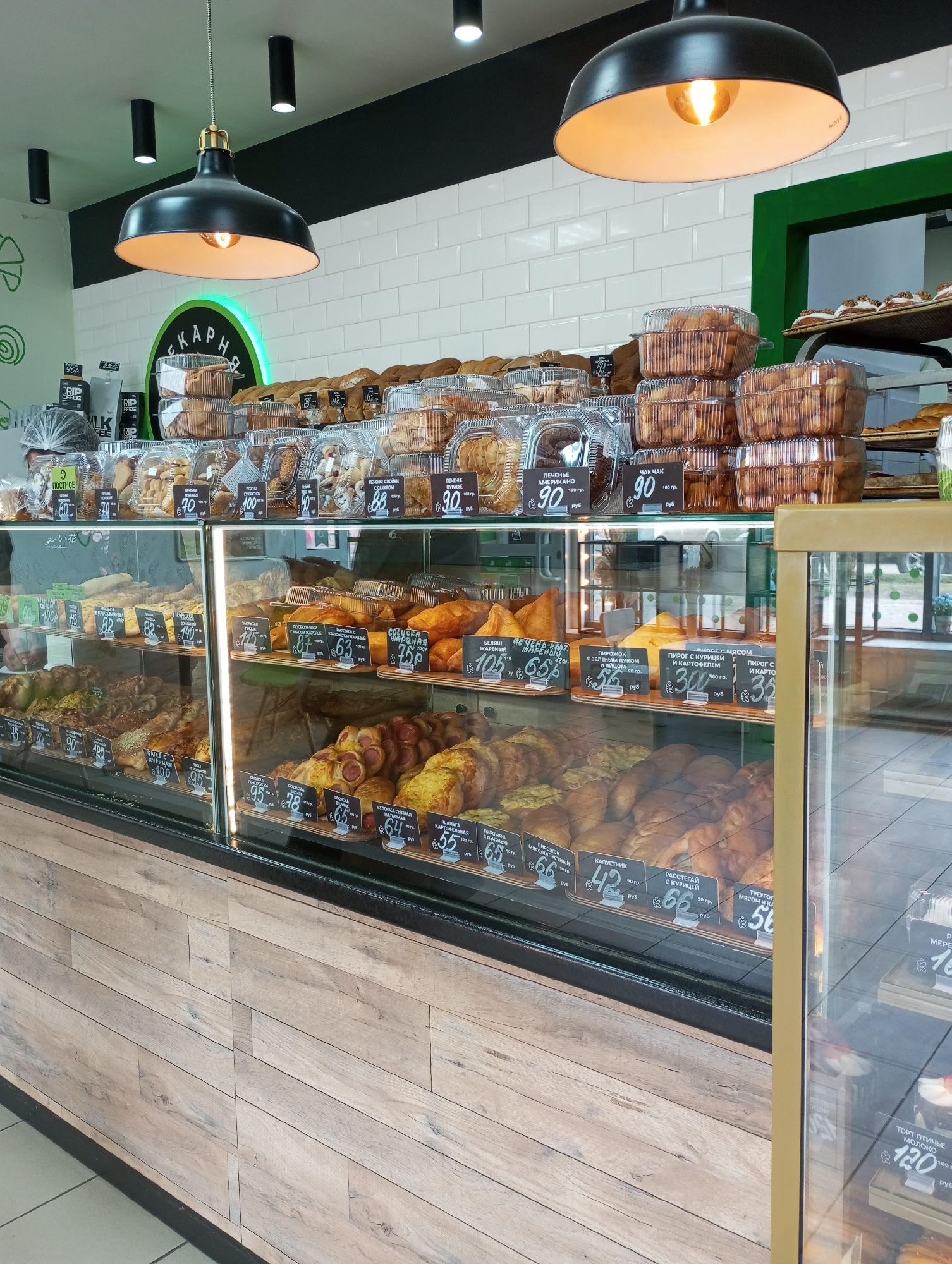 The image captures a bakery display case in Izhevsk, Russia. The case is filled with various baked goods, including packaged cookies, pastries, and pies, each labeled with its price in Russian rubles (руб) per weight in grams (гр). Prices range from 35 to 120 rubles. The display case is situated within a bakery, evident from the word "ПЕКАРНЯ" (bakery) on the wall, along with the green circle design. The case has a wood-like paneling on the lower section, and overhead lighting illuminates the goods. The black ceiling features track lighting fixtures, with two large pendant lamps hanging above the display case. The environment suggests a commercial setting with a focus on the sale of baked goods.