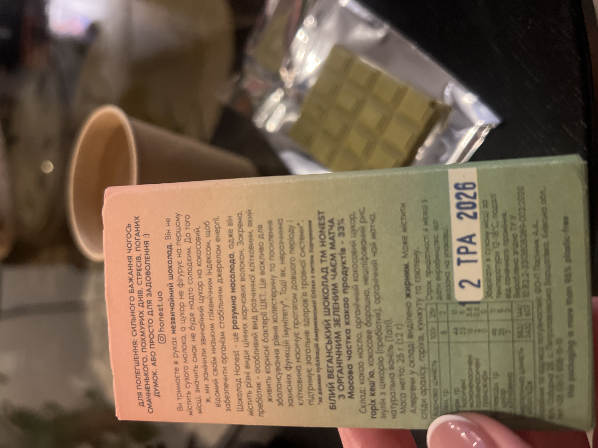 The image shows a close-up of a box of Honest brand vegan chocolate with matcha, held by a person's hand with a manicured fingernail. The text on the box is in Ukrainian and discusses the benefits of the chocolate, such as being a source of prebiotic fiber and not containing dairy or refined sugar. It also lists ingredients and nutritional information, with a best-by date of 2026. Behind the box, a disposable coffee cup is visible, and to the right, a piece of yellow chocolate is partially visible through clear plastic packaging. The background is out of focus and appears to be a dark surface, possibly a table. The text on the packaging also includes an address in Kyiv, Ukraine: "м. Київ, вул. Софіївська 25, с. Петропавлівська Борщагівка, Київська обл." which translates to "Kyiv city, Sofiivska St. 25, s. Petropavlivska Borshchahivka, Kyiv region."