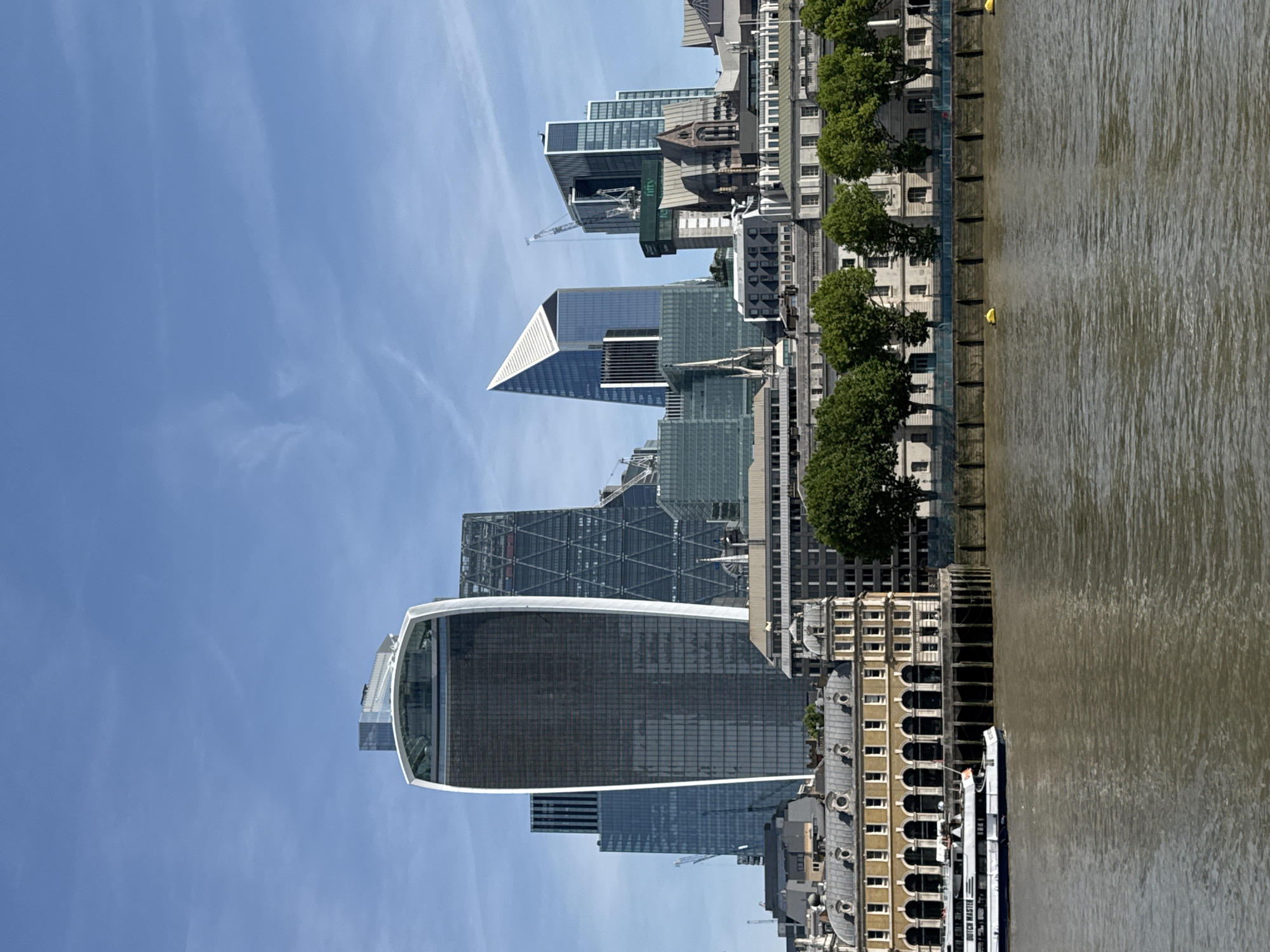 The image captures a view of the City of London skyline from across the River Thames. Key landmarks include the "Walkie-Talkie" building (20 Fenchurch Street), other skyscrapers with distinctive architectural designs, and a construction crane in the distance. A section of the riverfront is visible, with buildings and trees lining the bank. A boat is docked along the river. The sky is clear and blue.