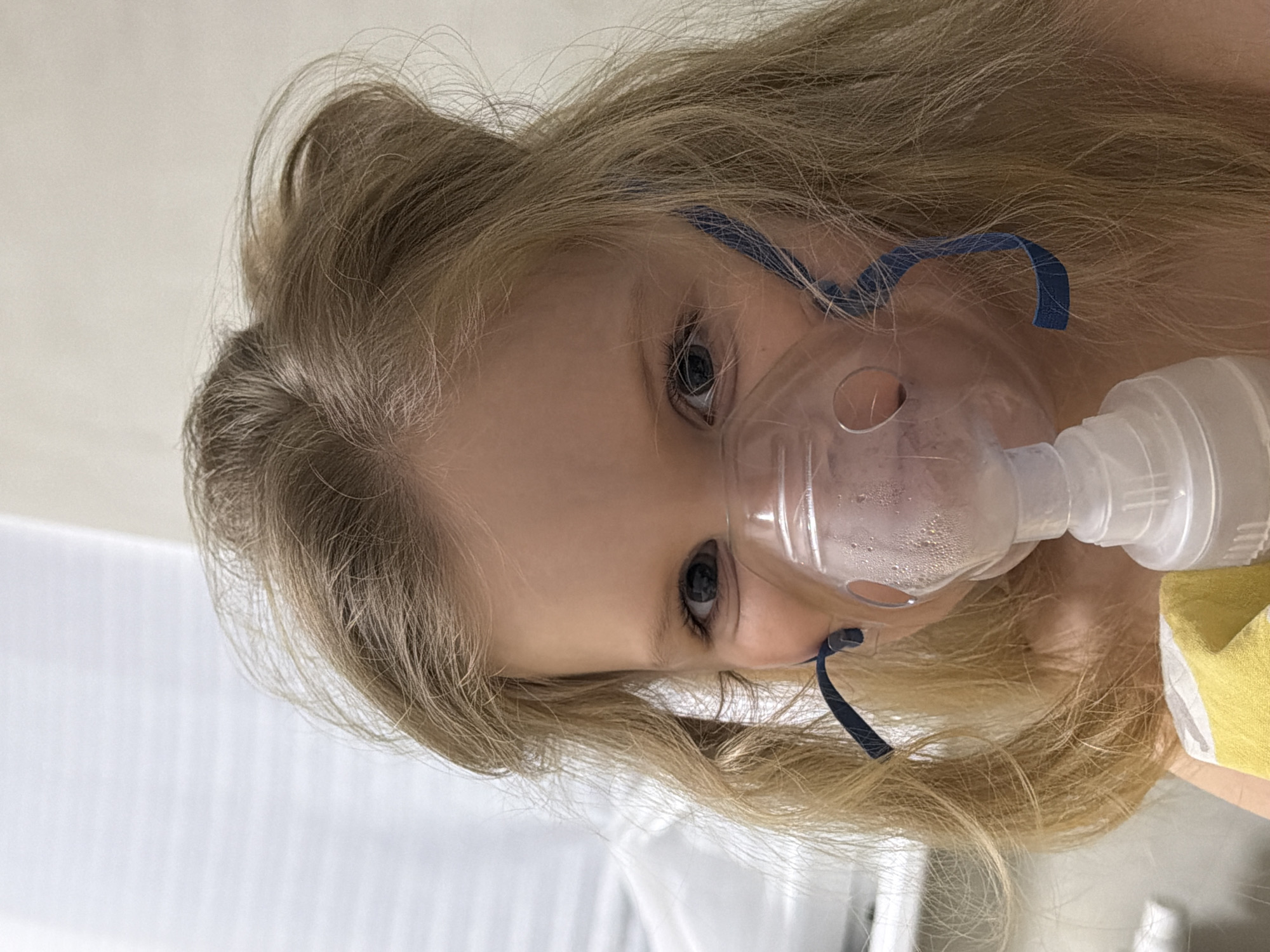 A young child with blonde hair is wearing a clear nebulizer mask with blue straps. The child's eyes are visible and appear to be looking towards the camera. The nebulizer is connected to a small, white plastic container. Part of a yellow and white patterned object is visible to the right of the child. The background is blurred and shows white surfaces. There is no verifiable information to confirm the location of Chaikovsky, Russia.