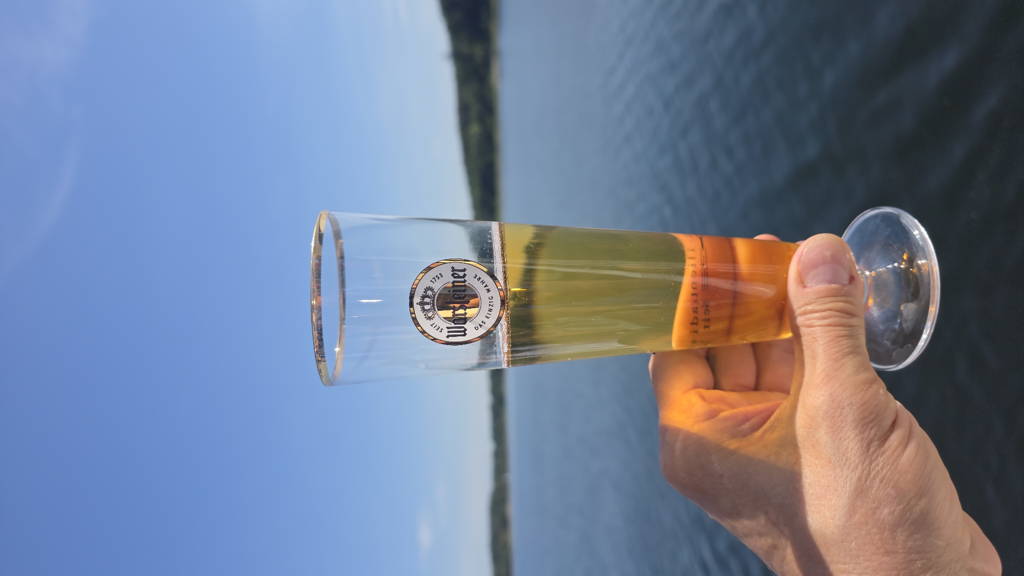 A glass of Warsteiner beer, with a golden rim and a logo featuring a crown and the text "Seit 1753 Warsteiner Das einzig wahre", is held up by a hand against a backdrop of a bright blue sky and a body of water, likely the Möhnesee in Germany, which is calm and reflects the sky. The beer is pale yellow with a small head of foam. There are two pieces of orange fruit submerged in the beer at the bottom of the glass. The sun is shining brightly, casting a warm glow on the hand and the glass. The horizon line is visible in the distance, with a faint outline of trees on the far shore. The overall scene evokes a sense of leisure and enjoyment on a clear day.