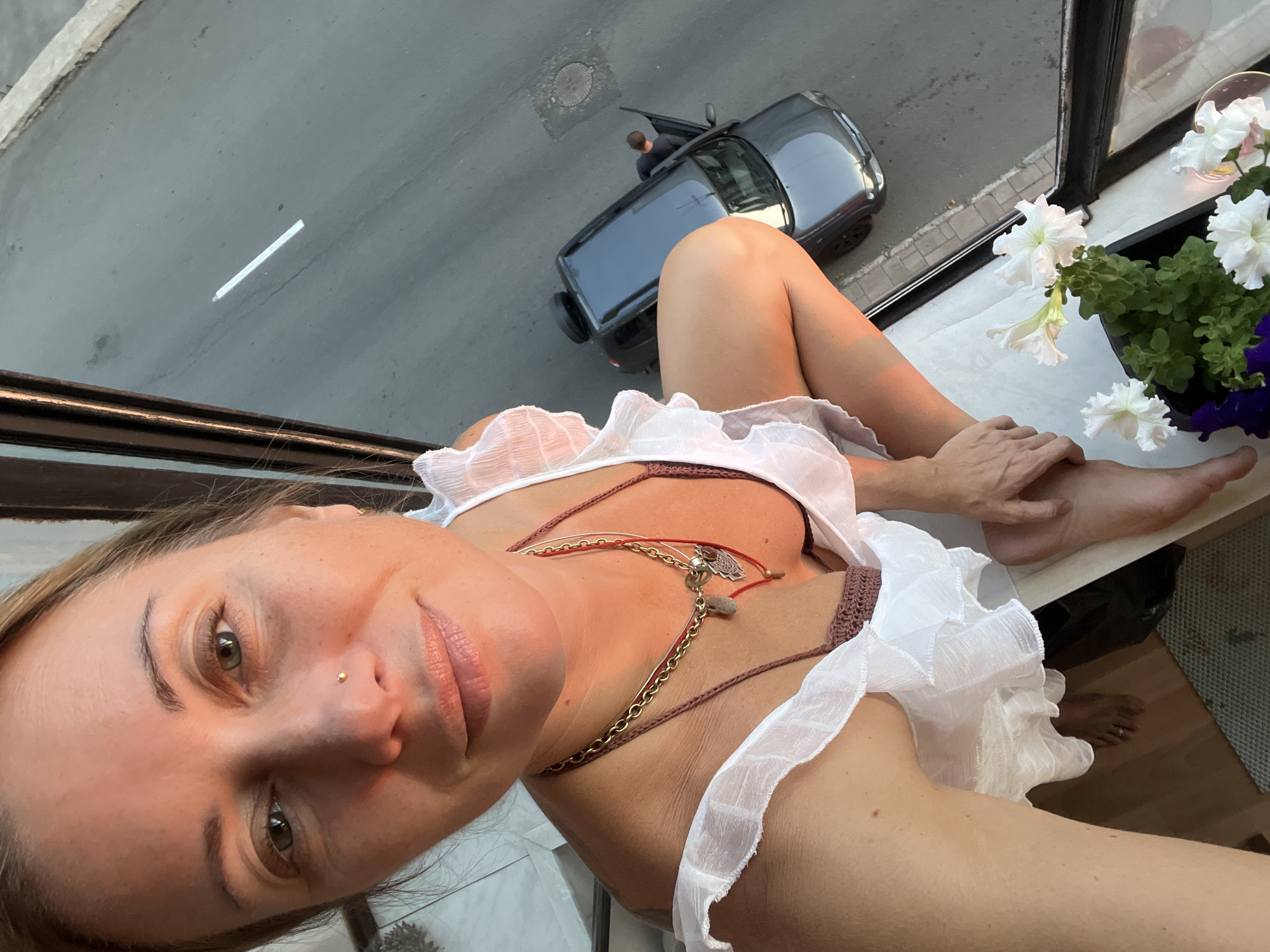 A woman is perched on a windowsill, looking down at a street. She is wearing a white, ruffled garment and a necklace. Her right leg is bent, and her left foot is visible. There are white flowers in a pot on the windowsill. Below, a dark-colored car is parked with its door open. A man in a dark shirt is standing next to the car. The scene appears to be set during the day, possibly late afternoon given the warm lighting. The background suggests an urban environment with a road and a manhole cover visible. The overall mood is relaxed and casual. There is no visible text in the image.