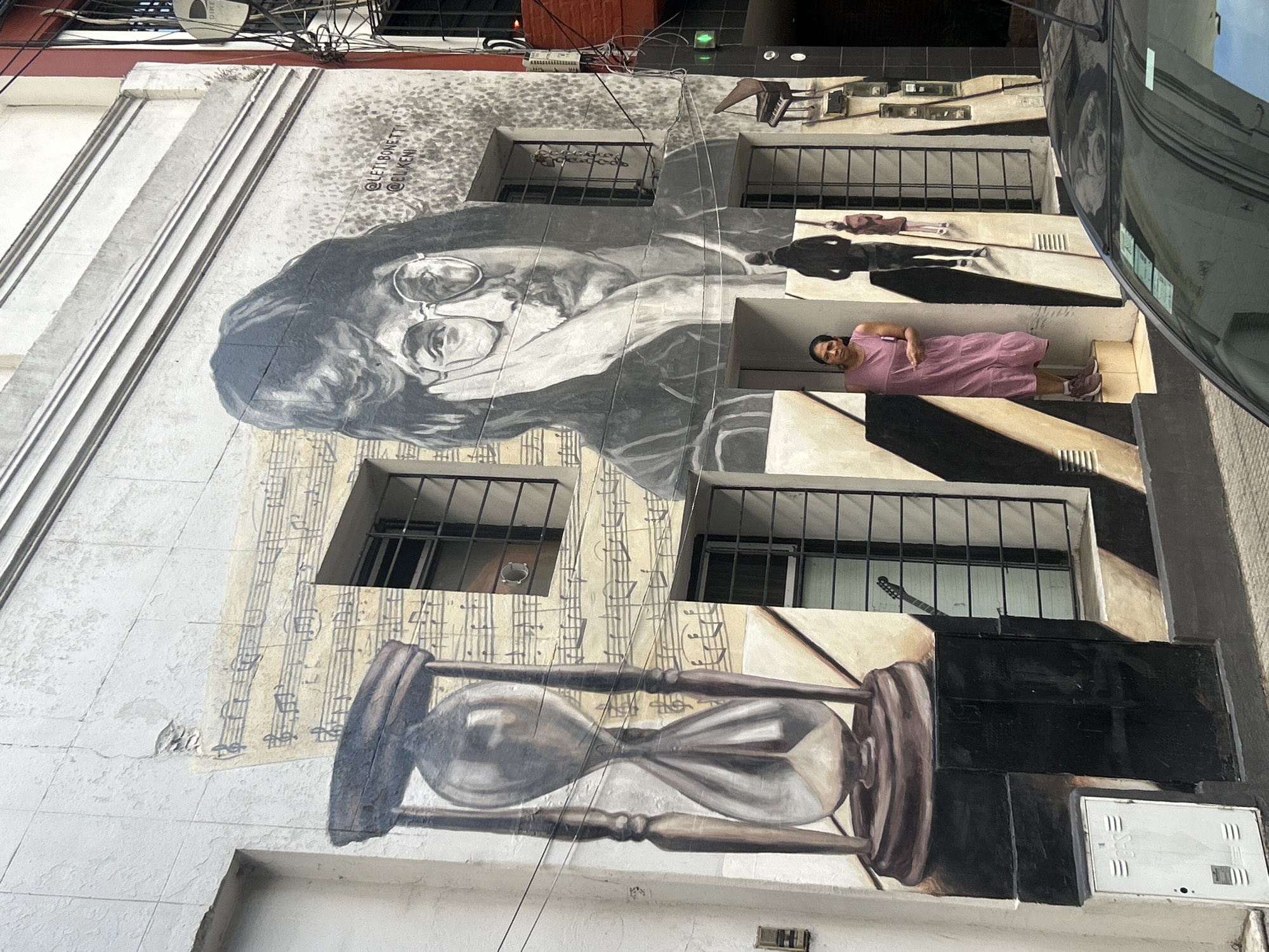This image shows a mural on a building in Buenos Aires, Argentina. The mural depicts a portrait of John Lennon with his iconic round glasses, musical notes, and an hourglass. Below John Lennon's face, an hourglass is depicted against musical note background. The mural interacts with the building's architecture; people are painted on ledges/window sills; woman in a pink dress, a silhouetted figure, and some other figure in the distance. The mural also interacts with the building's windows; a guitar appears to be behind bars. Cables and a piece of a car are visible in the frame.