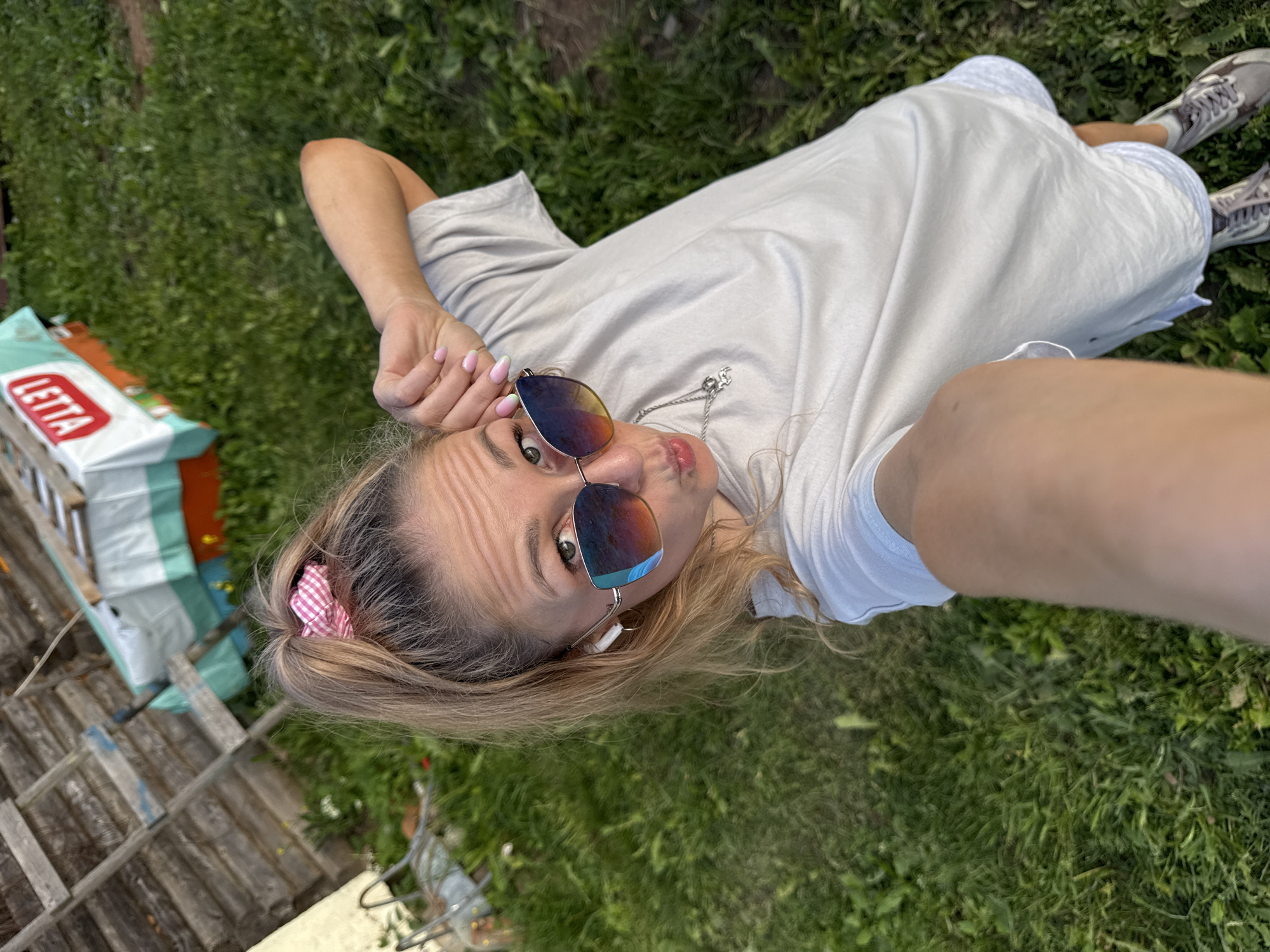 A young woman lies on her back on a grassy lawn, looking up at the camera. She has blonde hair pulled back with a pink and white checkered scrunchie. She is wearing a light gray t-shirt and white shorts, with gray athletic shoes. Sunglasses with mirrored lenses are perched on her nose, and she has her right hand near her face, fingers outstretched. Her lips are pursed. The scene is outdoors, with green grass and some wooden structures in the background. A white bag with the word "LETTA" in red lettering is visible to the left. The lighting suggests it is daytime, likely sunny, and the overall mood is relaxed and playful.