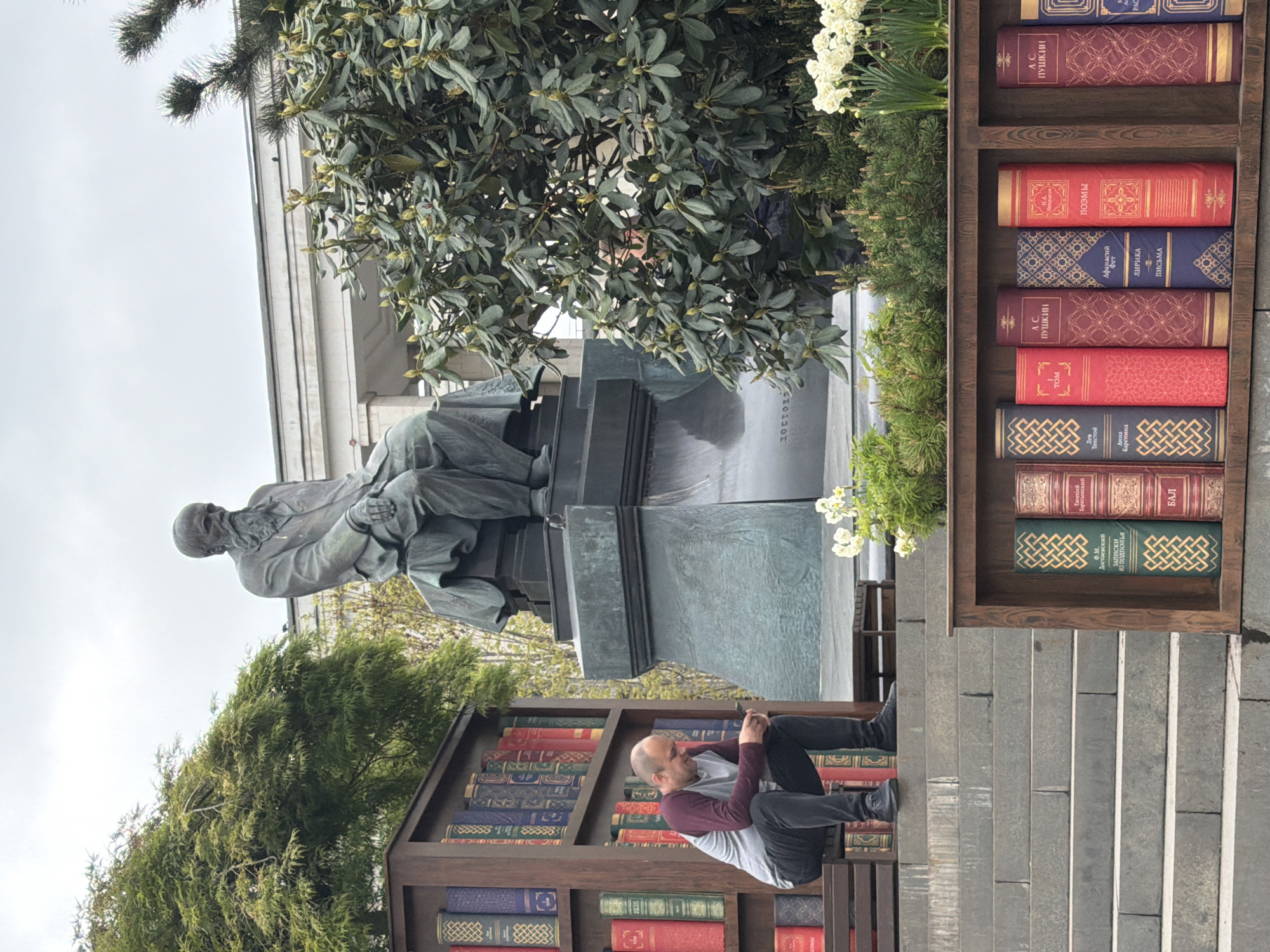 The photograph shows a public square in Moscow, Russia, notable for its literary theme. At the center of the image is a bronze statue of a seated man. It is positioned atop a stone pedestal. The inscription "ДОСТОЕВ" is seen on the stone base. The statue is surrounded by greenery, including trees and flowering plants.
To the right and left of the statue are large, decorative bookcases constructed of wood. They feature spines of Russian literature books. One bookcase is occupied by a person; a man wearing casual clothing is sitting on a shelf. He is looking at his smartphone.
The setting seems to be an urban public space, possibly a park or a plaza. The sky is overcast. The foreground of the image includes paved steps leading up to the scene. The bookcases are part of an open-air installation or exhibit.