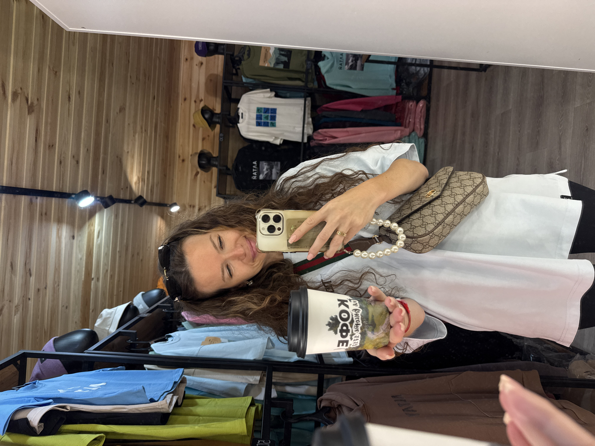 A woman is taking a selfie in a clothing store. She is wearing a white t-shirt and black pants. She is holding a coffee cup in her left hand and her phone in her right hand. The coffee cup has a picture of a tree and the word "КОФЕ" (coffee in Russian) on it. She is also wearing a pearl necklace and a Gucci bag. The store is filled with clothes, hats, and other merchandise. The walls and ceiling are made of wood. The floor is made of laminate. The scene appears to be in T/B Seminskii Pereval, Russia.