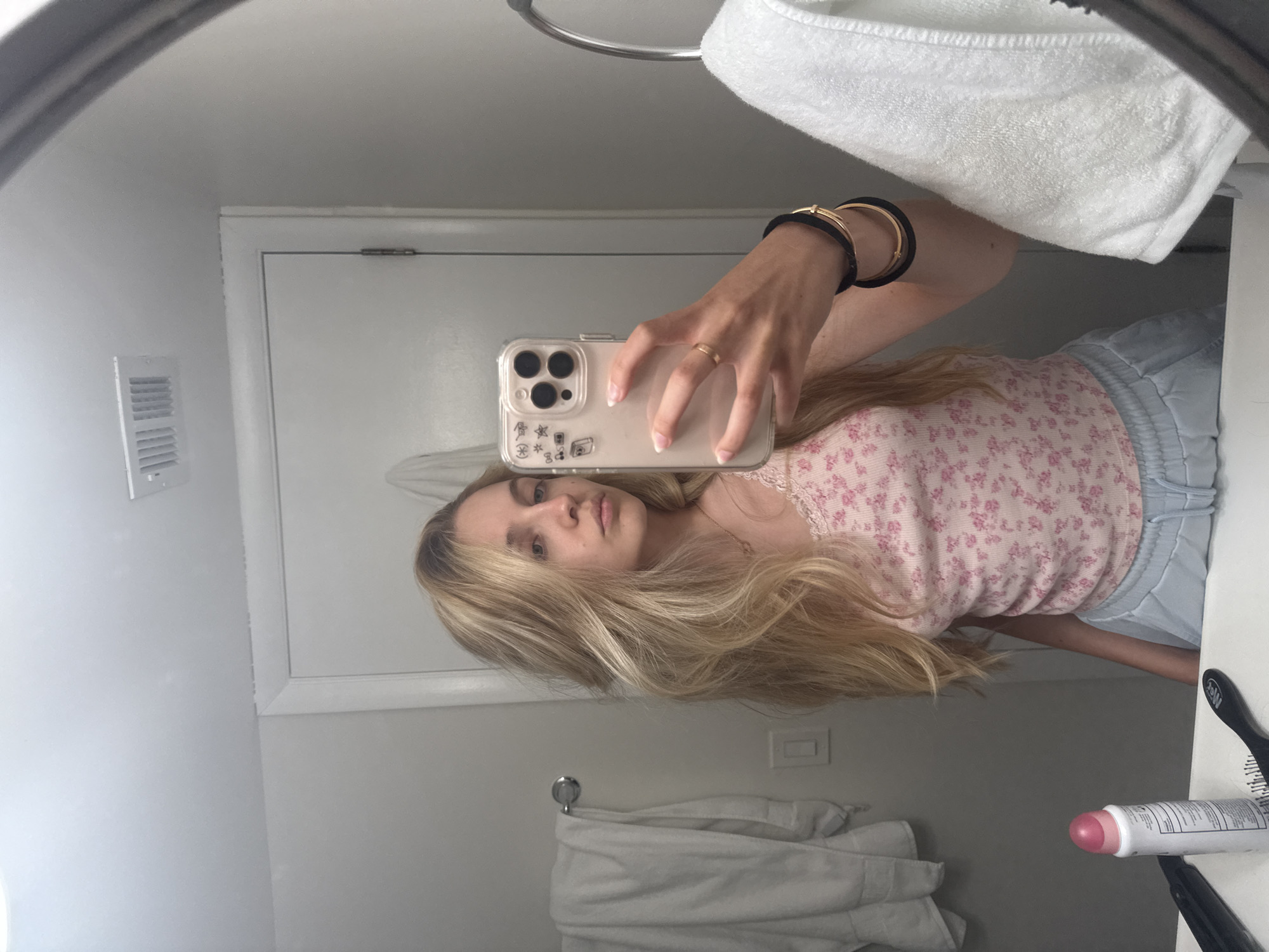 A young woman with blonde hair tied back takes a mirror selfie in a bathroom. She is wearing a pink floral tank top and light blue sweatpants. Her left hand is holding her phone, which has a clear case decorated with stickers. She is also wearing a gold ring and black and gold bracelets on her left wrist. In the background, a white towel hangs on a rack to the left, and another white towel is visible to the right. A white door is in the background. The scene appears to be in a residential bathroom.