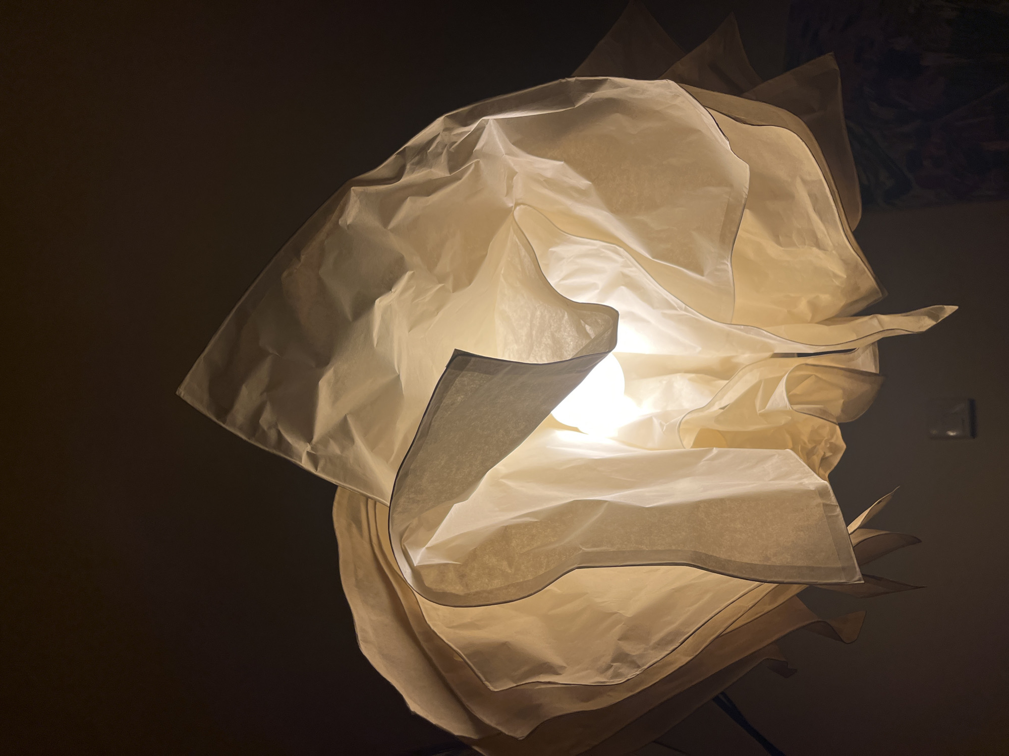 The image shows a lamp with a lampshade made of layered, translucent material, likely paper or a similar lightweight fabric. The light source is inside the shade, illuminating the material and creating a soft, diffused glow. The background is dark and indistinct, suggesting the lamp is in a dimly lit room. The lamp is not identifiable as to the location in the city of Barcelona, Spain, and I cannot place it there.
