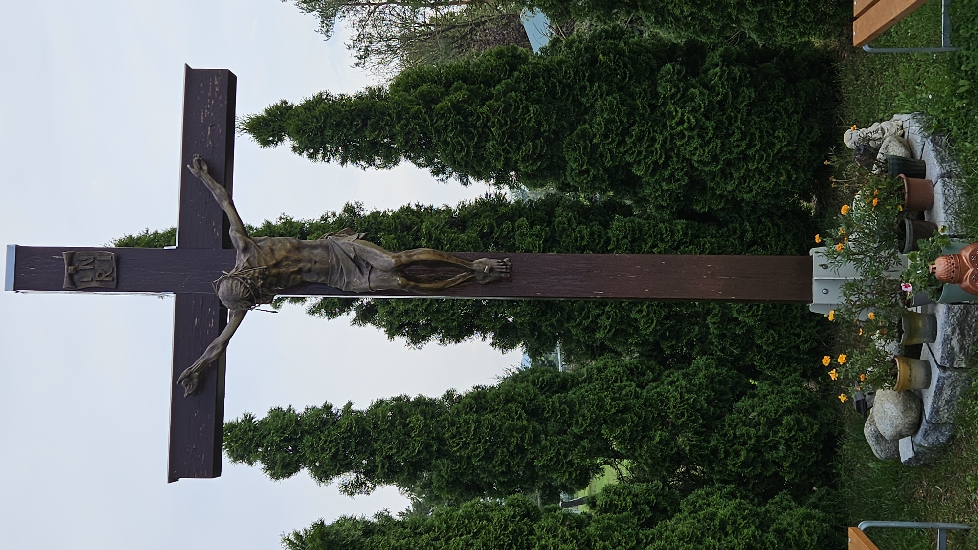 A large, dark brown wooden cross with a bronzed statue of Jesus Christ is positioned in front of a backdrop of lush green cypress trees under an overcast sky. The statue depicts Jesus with a crown of thorns and a loincloth, arms outstretched. Above Jesus' head, the inscription "INRI" is visible on the cross. At the base of the cross, a circular stone structure is adorned with several potted plants, some featuring small orange flowers, and a few large stones. A small angelic statue and an ornate lantern are also present. The scene appears to be outdoors, possibly in a garden or a memorial site. The location in the city of Prešov, Slovakia is not discernible from the image.
