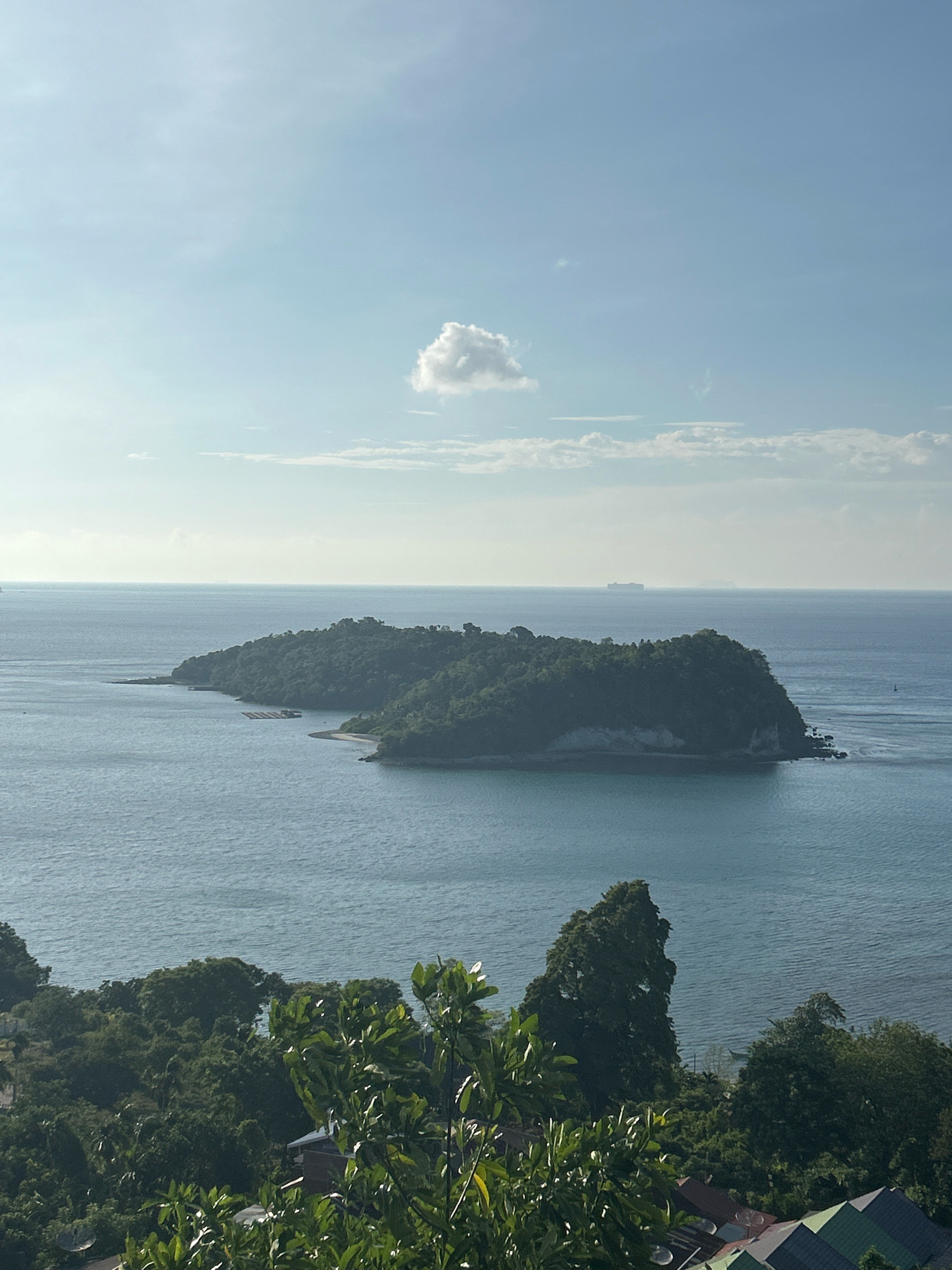 A panoramic view captures the serene seascape and lush green islands of Sabang, Indonesia, under a partly cloudy sky. The image showcases a large, verdant island in the midground, characterized by its densely forested slopes and rocky outcrops. In the distance, several ships are visible on the horizon, suggesting active maritime traffic. The foreground is dominated by a dense canopy of tropical trees, with hints of rooftops peeking through, indicating a populated area on higher ground overlooking the sea. The water is a calm, deep blue, with gentle ripples reflecting the soft light of the sky. The overall impression is one of natural beauty and tranquil coastal living.
