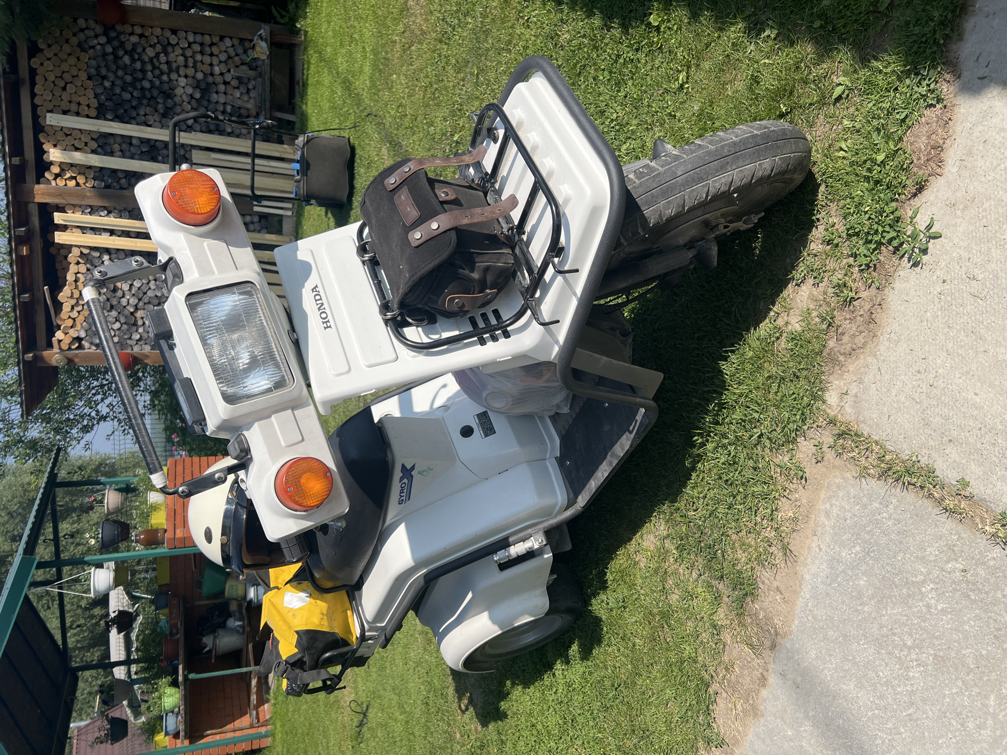 A white Honda Gyro X scooter is parked on a grassy lawn in front of a wooden pile of firewood and a brick structure. The scooter has a black seat, a yellow bag attached to the rear, and a black saddlebag on the front rack. A white helmet is placed on the seat. The background shows a green canopy structure with hanging flower pots and trees. The ground is covered in green grass and a concrete path.