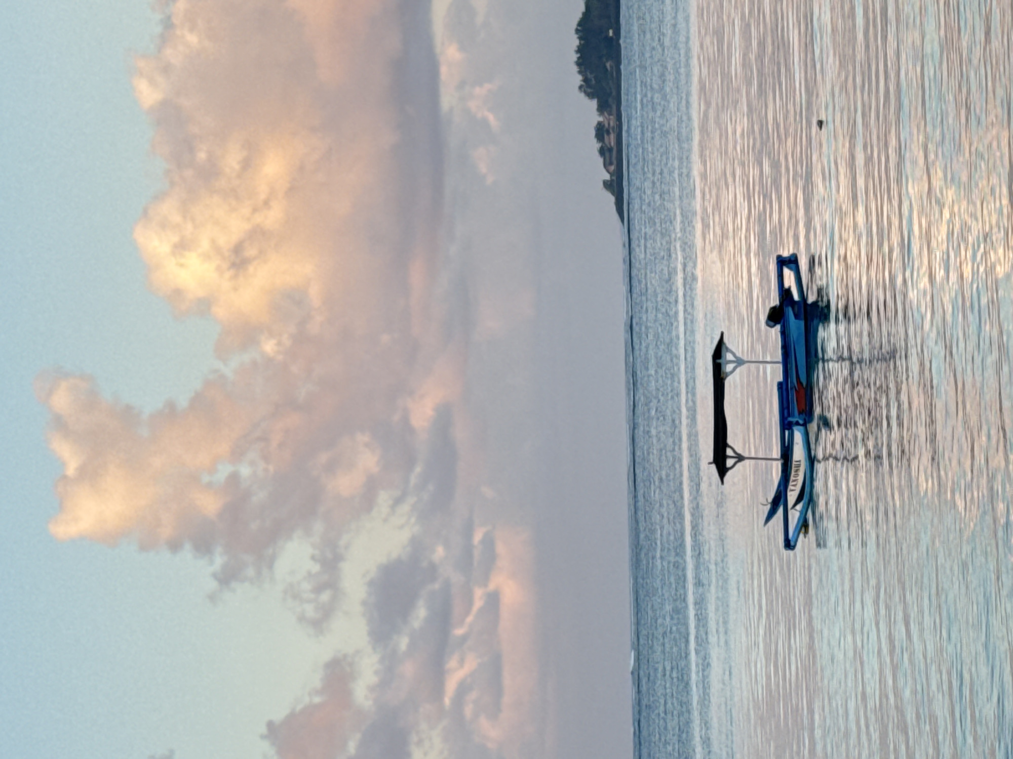 A small, blue and white outrigger boat with a dark canopy is anchored in calm, shallow ocean waters. The boat has a name, "VANUATU", written in white on its side. In the background, a distant, green island with buildings and trees is visible. The sky is filled with soft, pastel-colored clouds, suggesting either dawn or dusk. The scene is serene and peaceful, with gentle ripples on the water reflecting the sky's colors. The location is likely a coastal area near Denpasar, Indonesia, known for its beautiful beaches and tranquil waters.