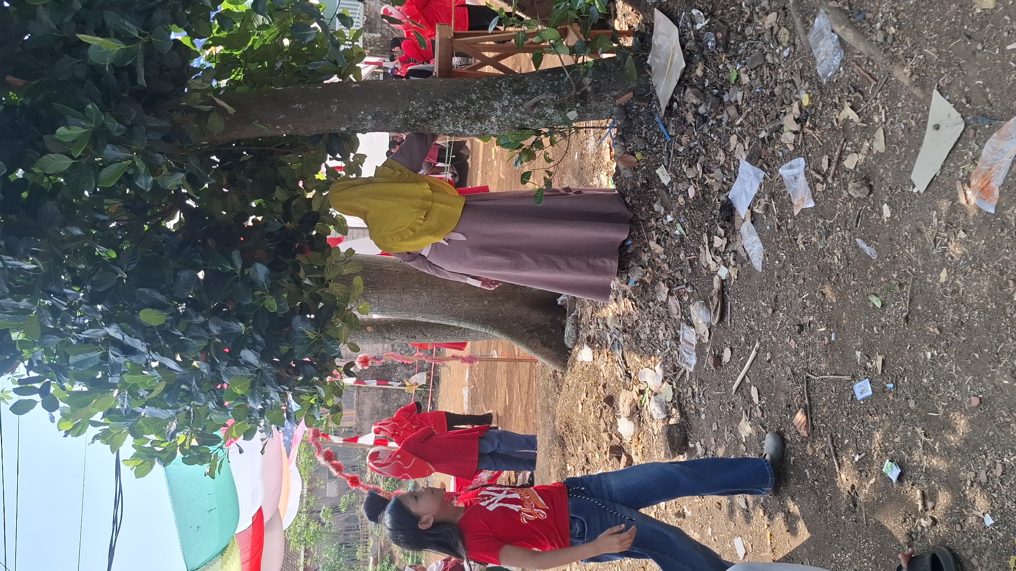 In Cilaku, Indonesia, an outdoor scene unfolds beneath the shade of a large tree. A young girl in a red shirt and blue jeans stands on the left, looking to her right. Further in the background, several individuals wearing red outfits and headscarves are visible, some appearing to participate in an event. A woman in a long, muted-colored dress and a yellow headscarf leans against a tree trunk. The ground is covered with dirt, scattered leaves, and numerous pieces of litter, including plastic bags and wrappers. Decorative banners, possibly red and white, are strung across the area, suggesting a festive occasion. The environment is somewhat unkempt, with visible debris on the ground.