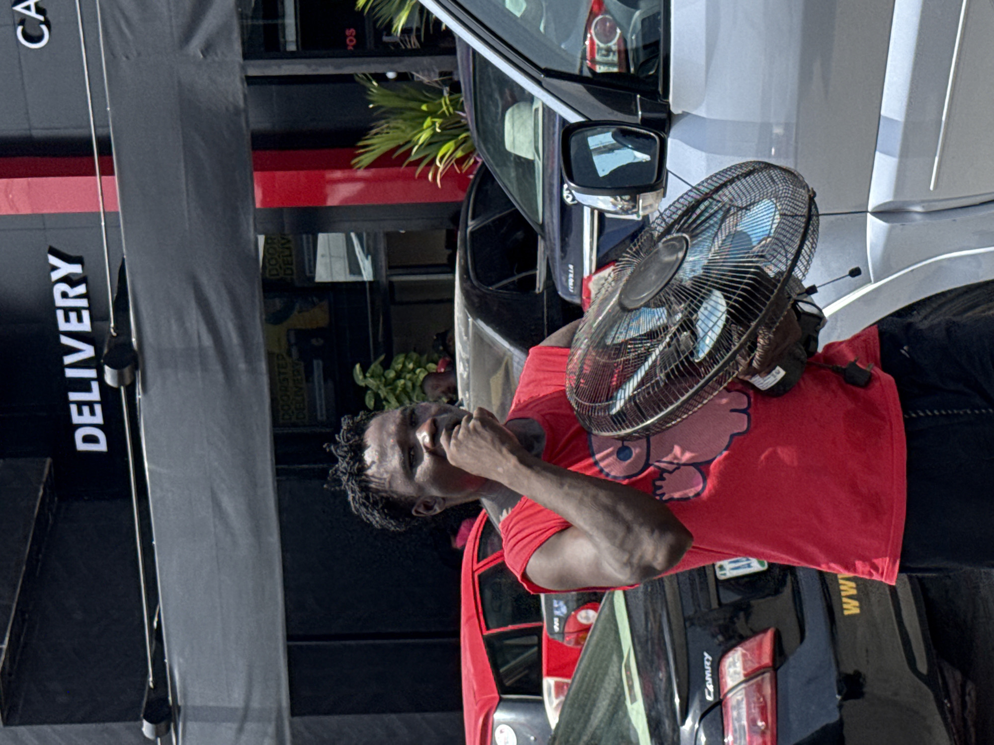 In Ilado, Nigeria, a man wearing a red t-shirt stands next to parked cars, holding a black fan to his face. The background includes a building with "DELIVERY" written vertically on the side.