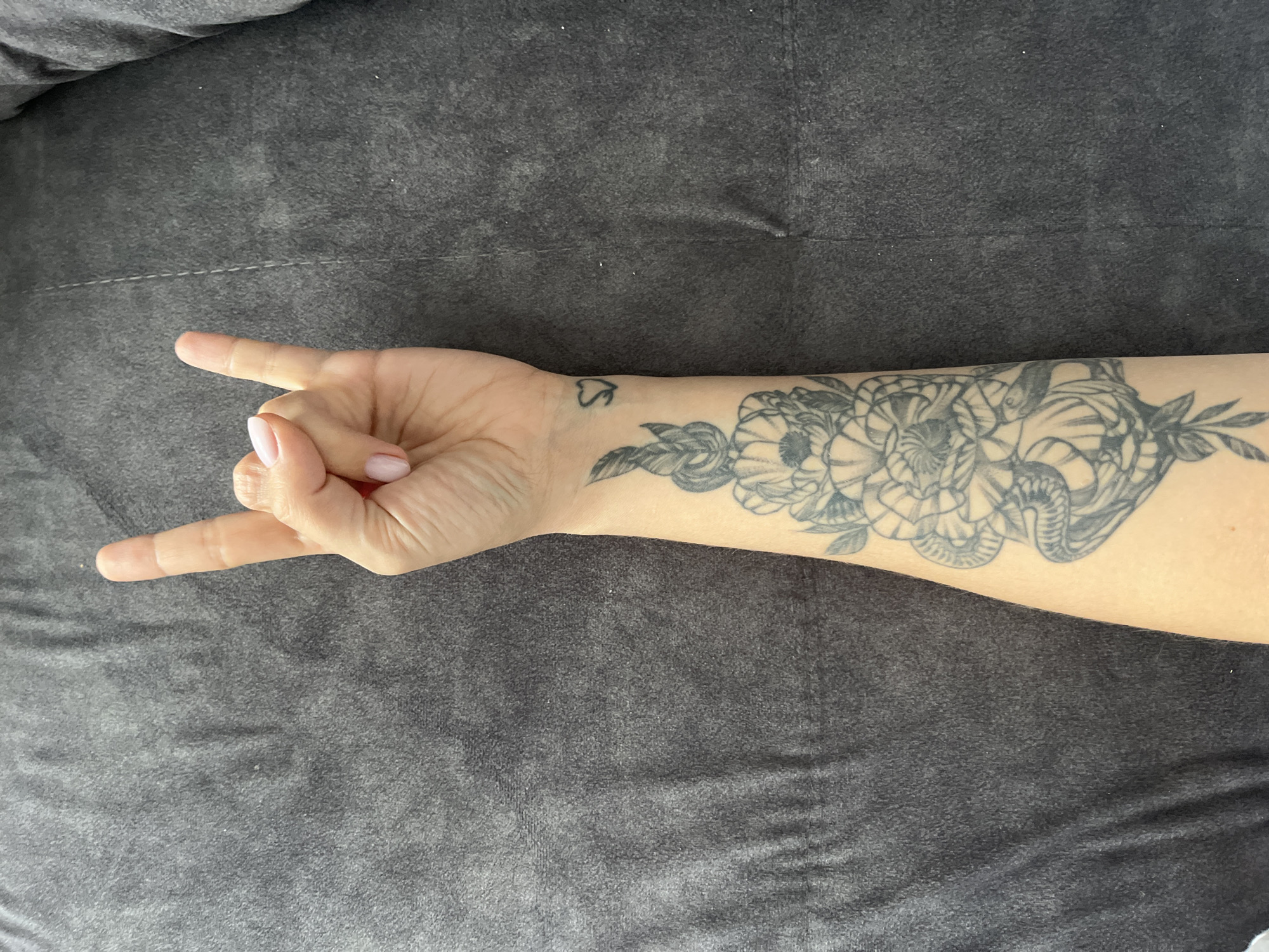 A human arm and hand is making a "rock on" gesture. The arm has a floral tattoo covering the forearm and a small symbol tattoo on the wrist. The background appears to be a gray fabric. There is no evidence indicating that the image was taken in Chaikovsky, Russia.